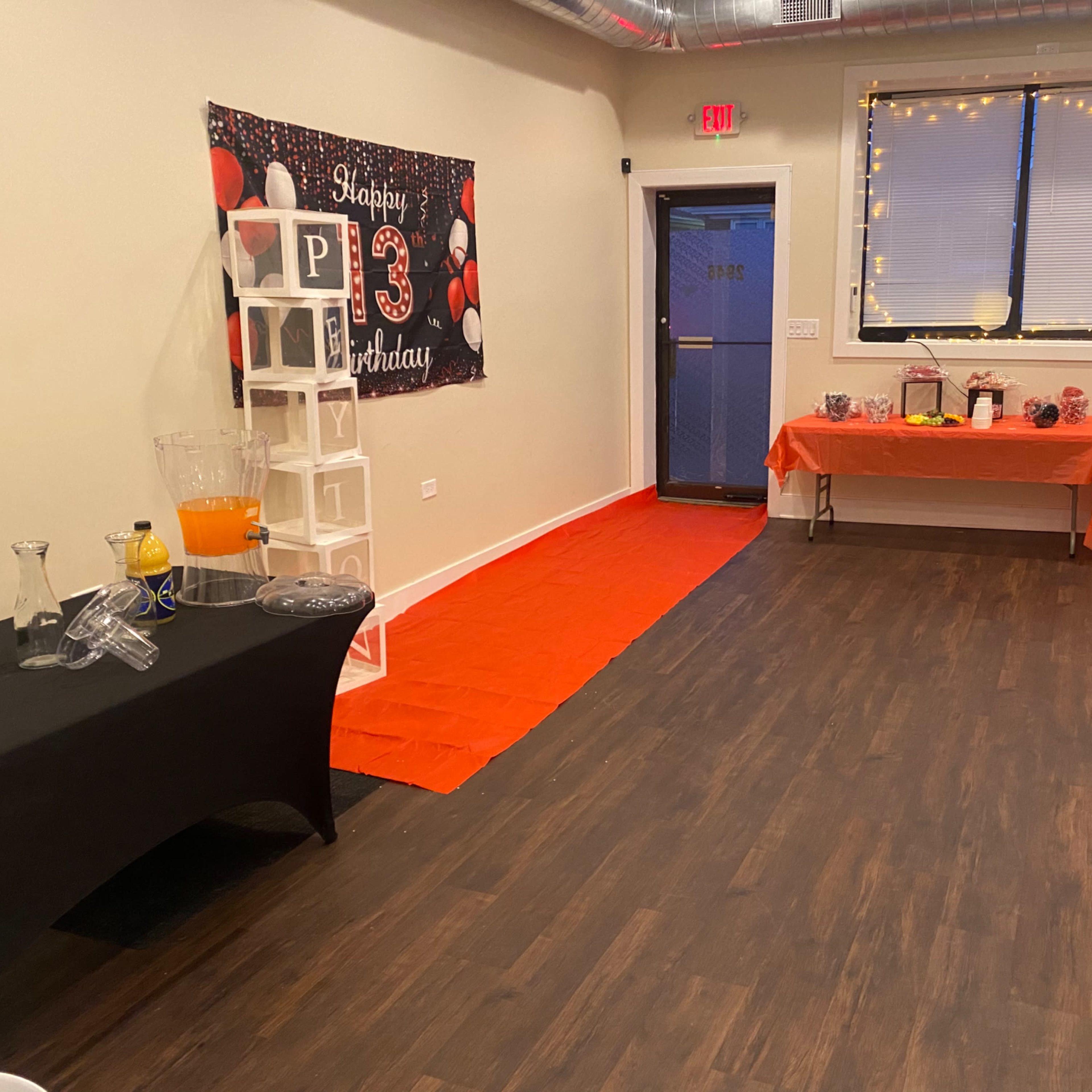 The interior of a party room features tables covered in black cloth, a decorative "Happy 13th Birthday" banner, and a bright orange carpet leading to a doorway.