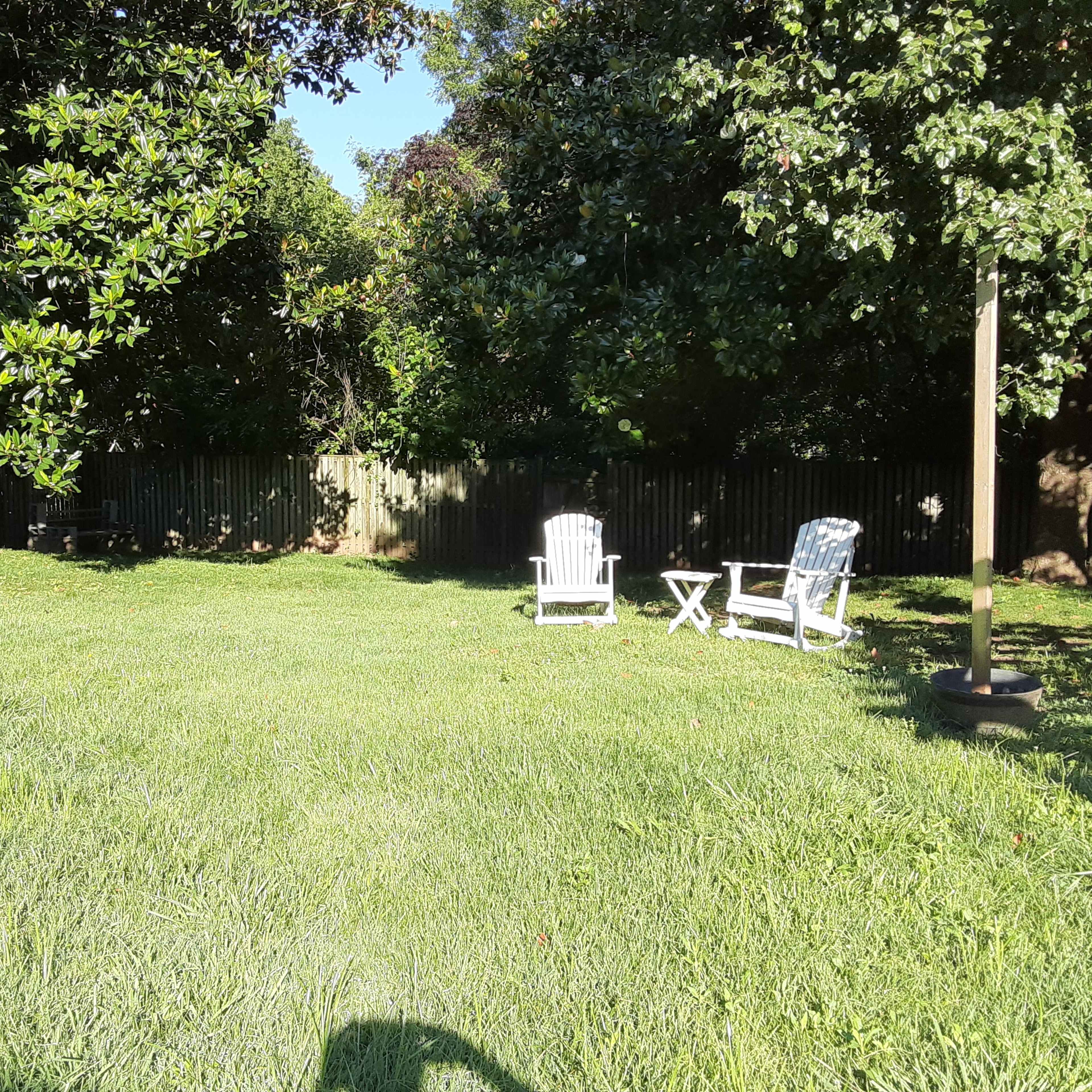 A grassy backyard features two white chairs and a small table under trees.