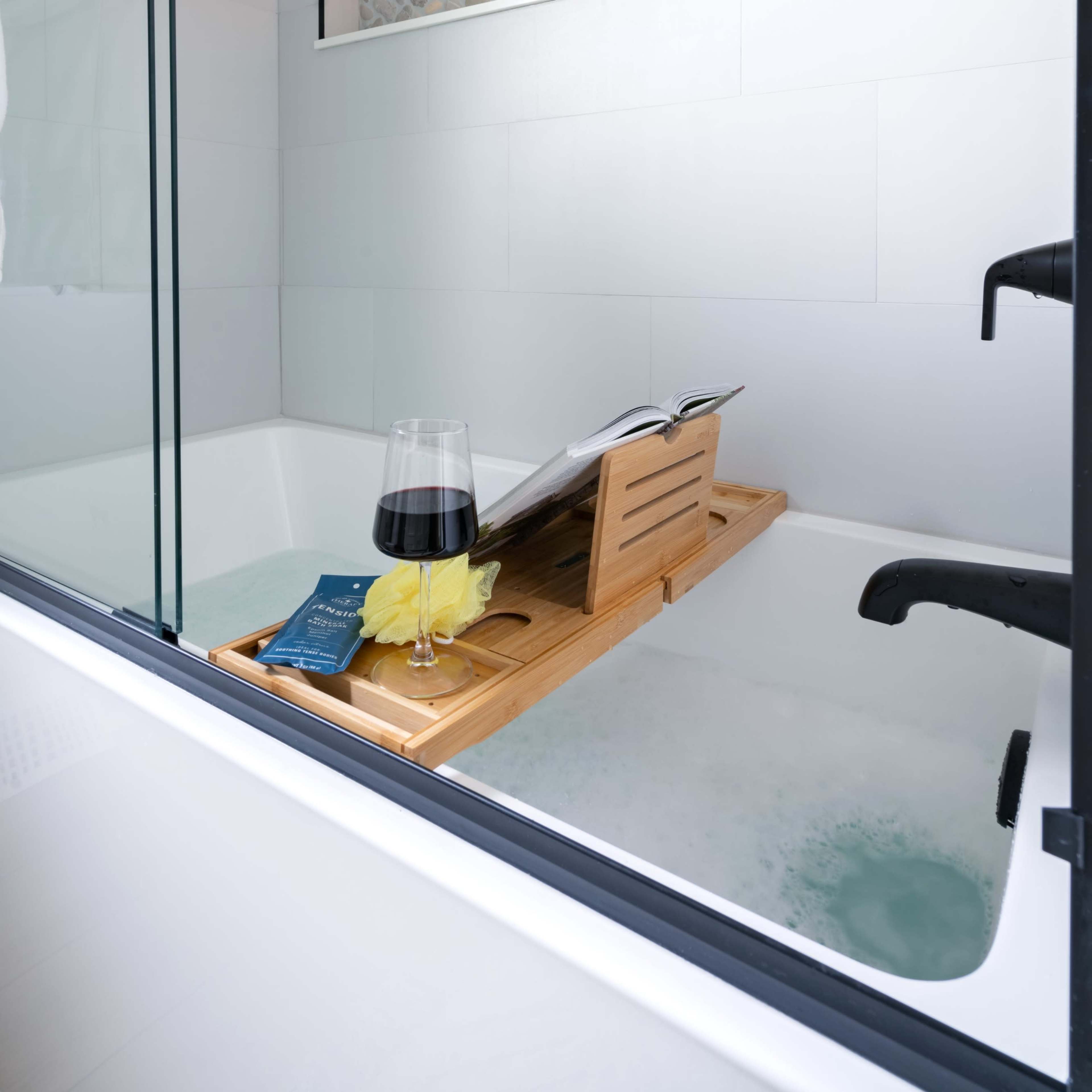 A wooden bath tray holding a book and a glass of wine rests on the edge of a filled bathtub with a decorative stone shelf illuminated in the background.