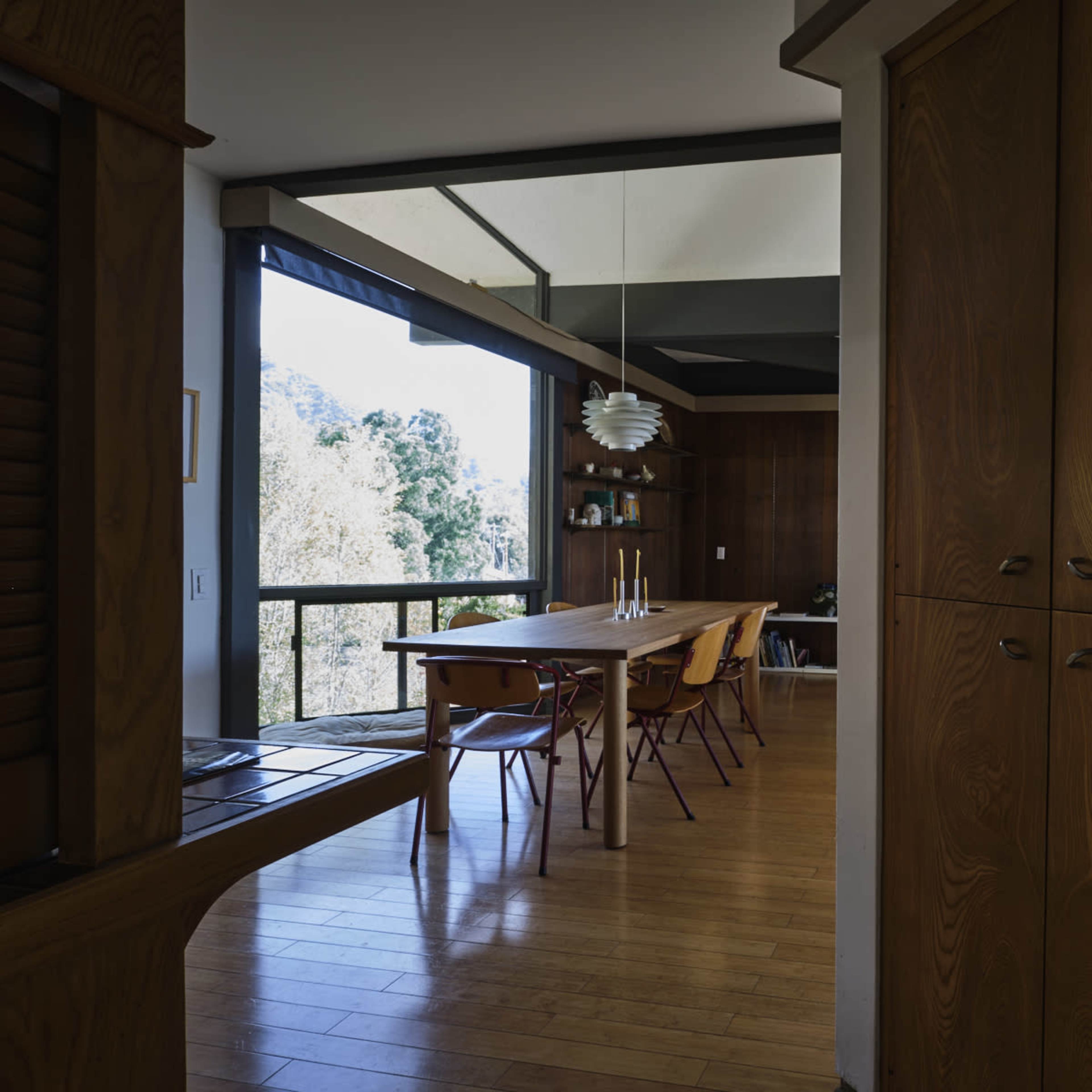 A modern dining area featuring a long table surrounded by chairs, with large windows providing a view of greenery outside.