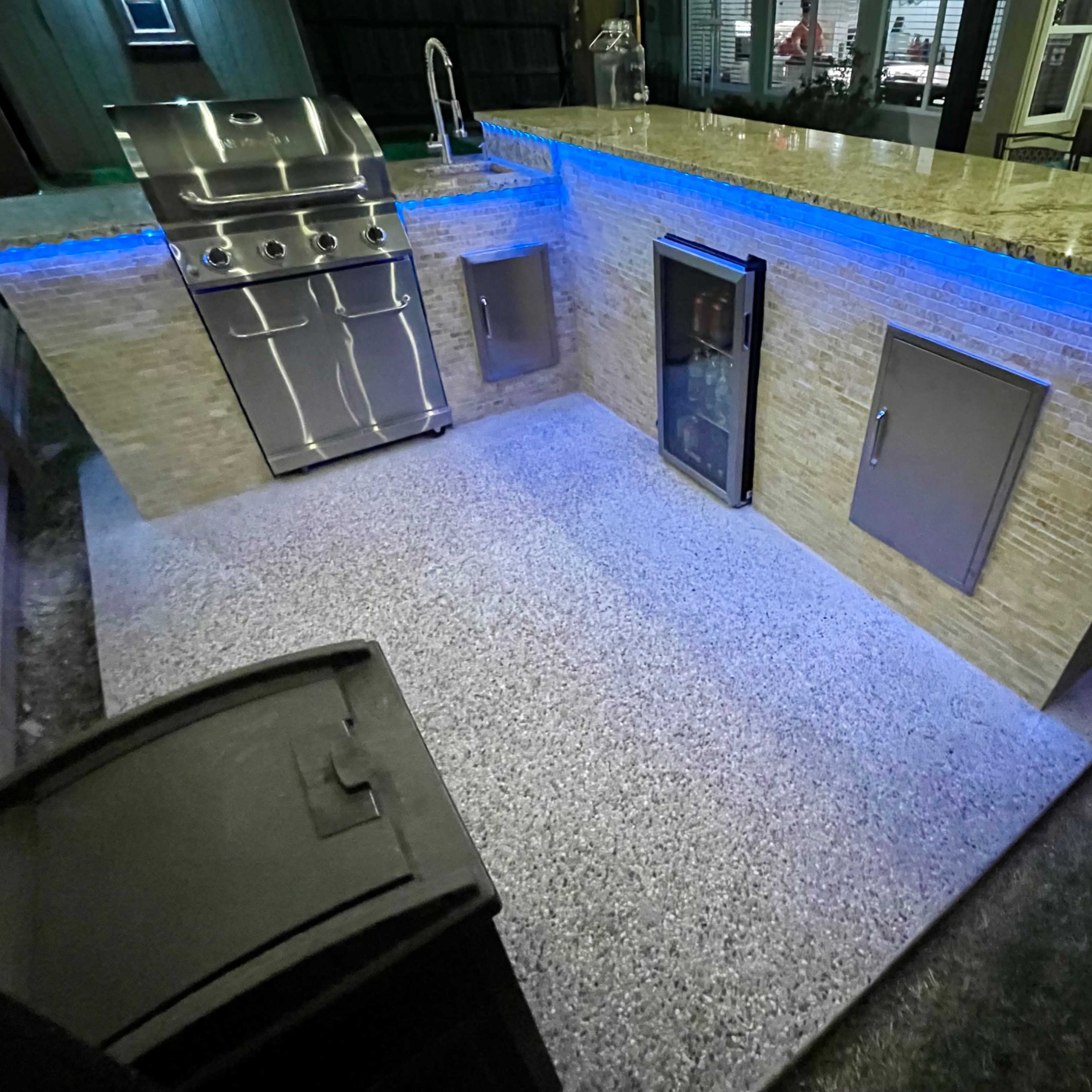 An outdoor kitchen area featuring a stainless steel grill, a refrigerator, and a granite countertop illuminated by blue lighting.
