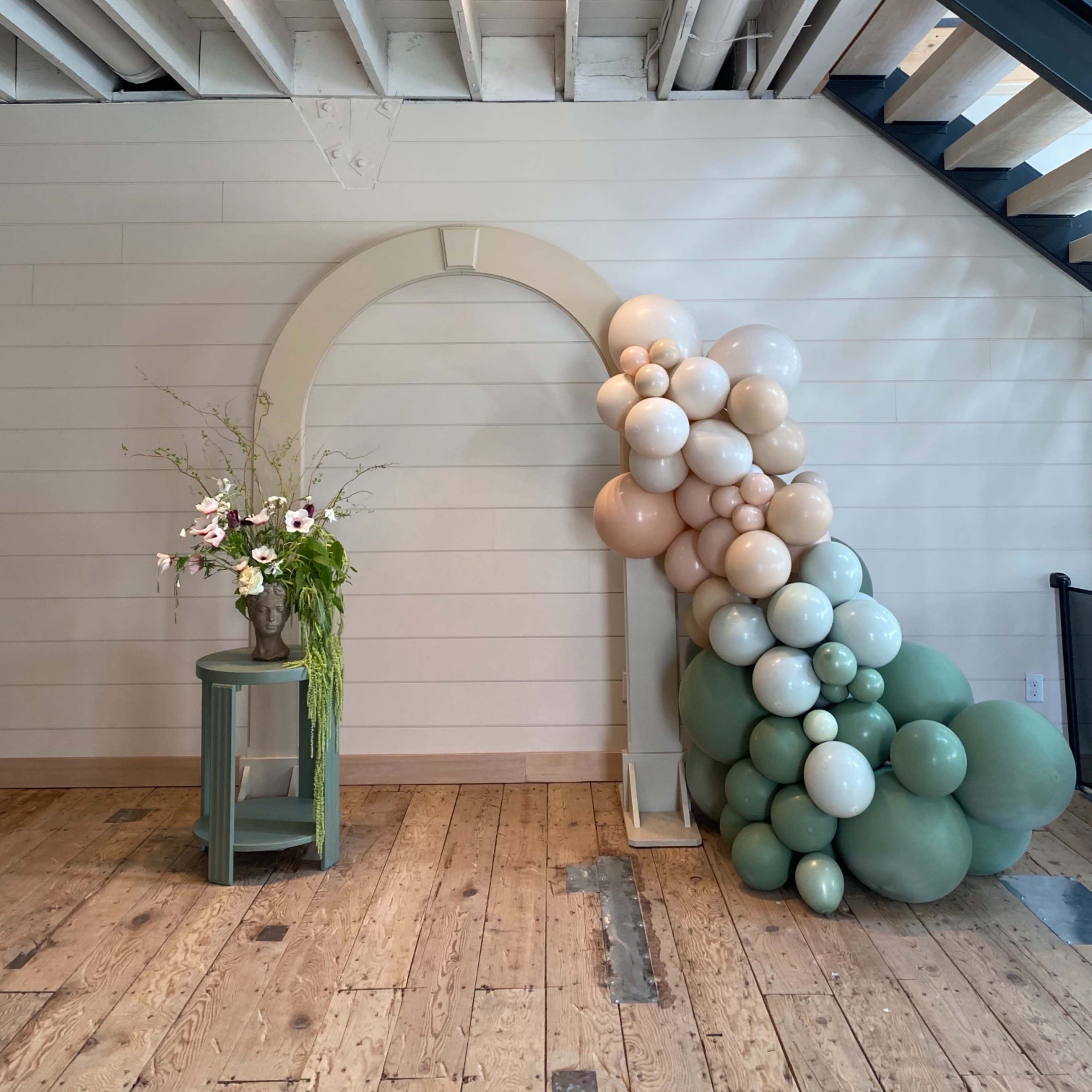 A decorative arch made of balloons in neutral tones stands next to a green side table with a floral arrangement, against a backdrop of wooden flooring and a staircase.