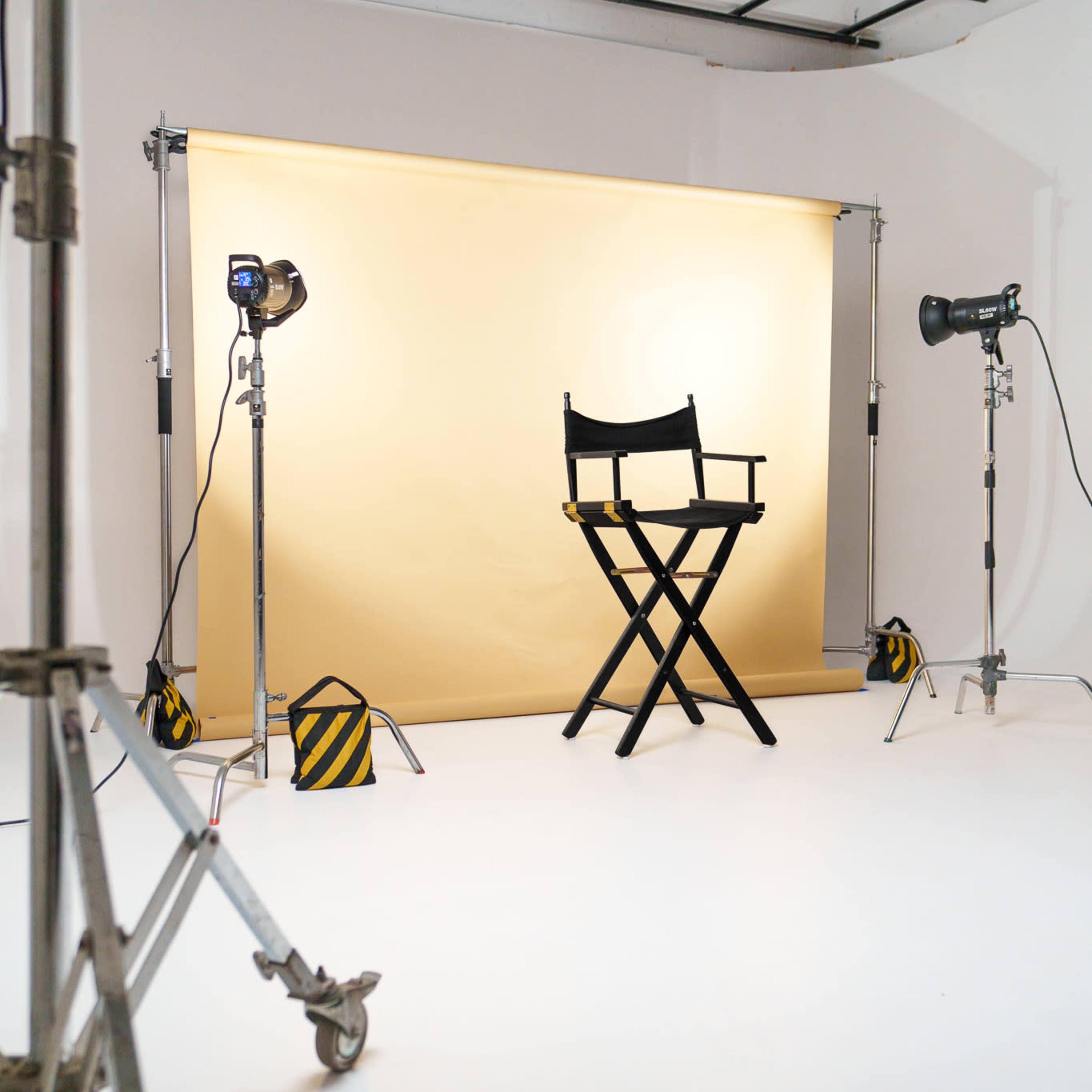 A photography studio features a folding director's chair in front of a yellow backdrop, surrounded by lighting equipment on stands.