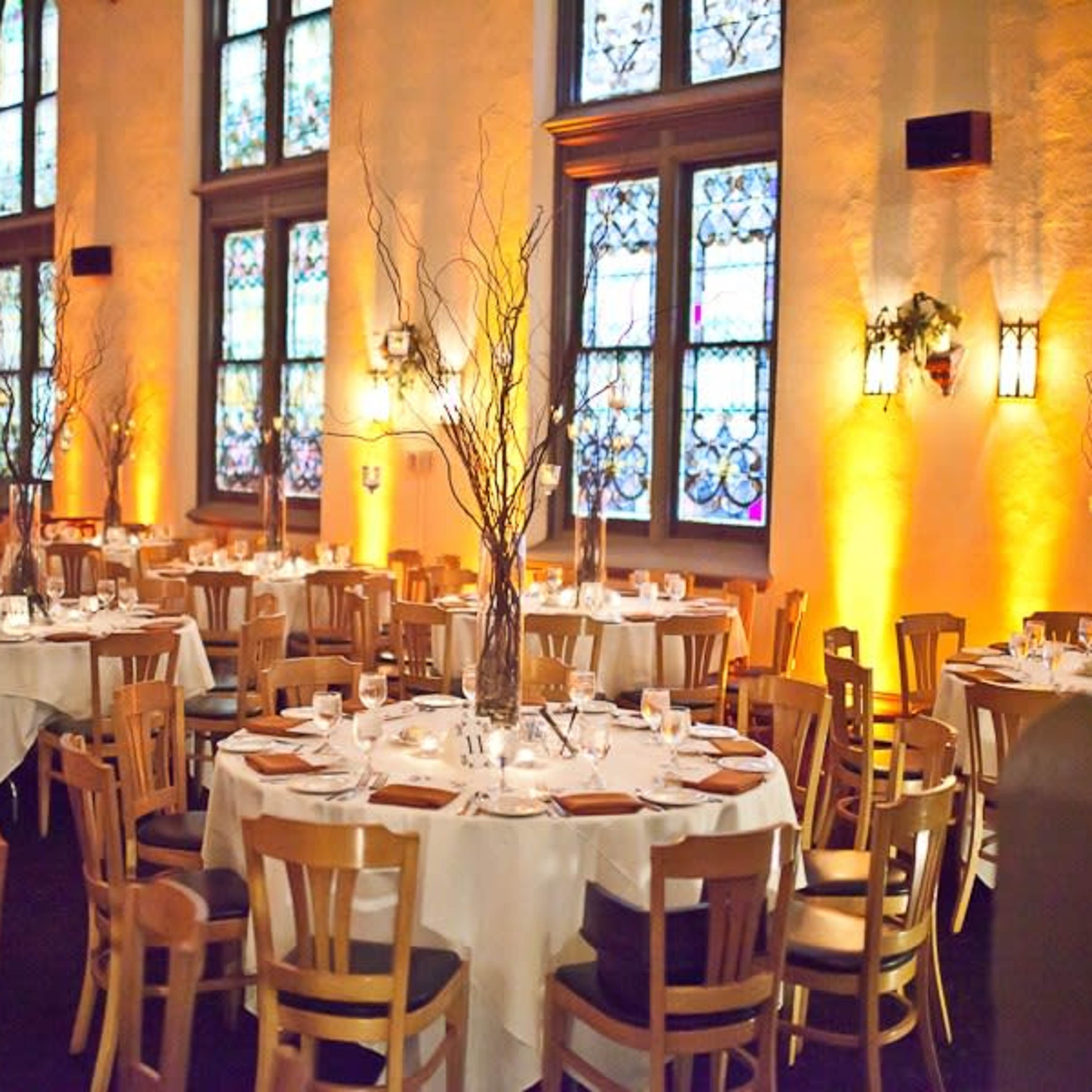 A dining area with round tables set for a meal, adorned with white tablecloths and central decorations, illuminated by ambient lighting and surrounded by stained glass windows.