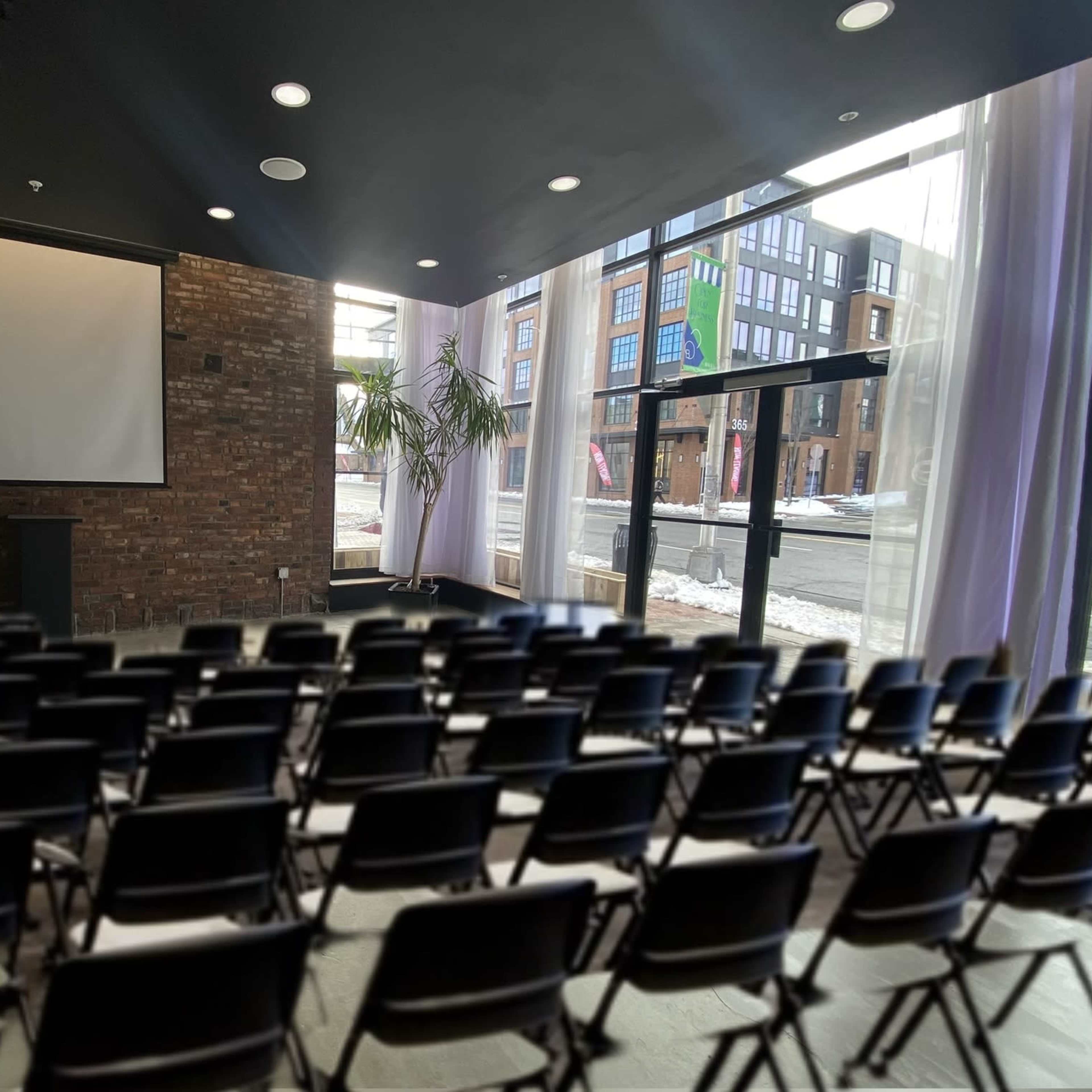 A meeting room with rows of black chairs facing a large projection screen, flanked by large windows.