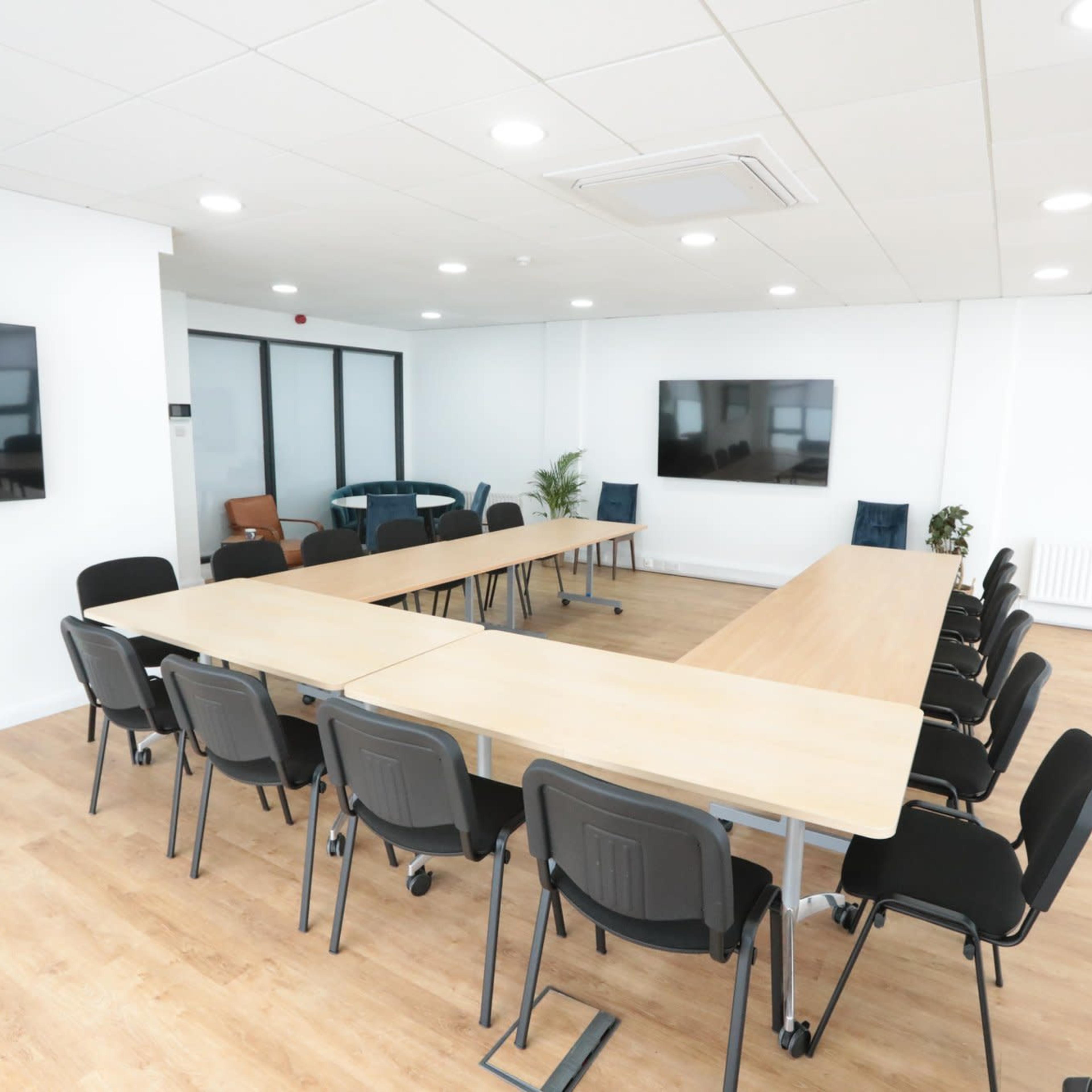 A large conference room features a long rectangular table surrounded by black chairs, with two screens mounted on the white walls.