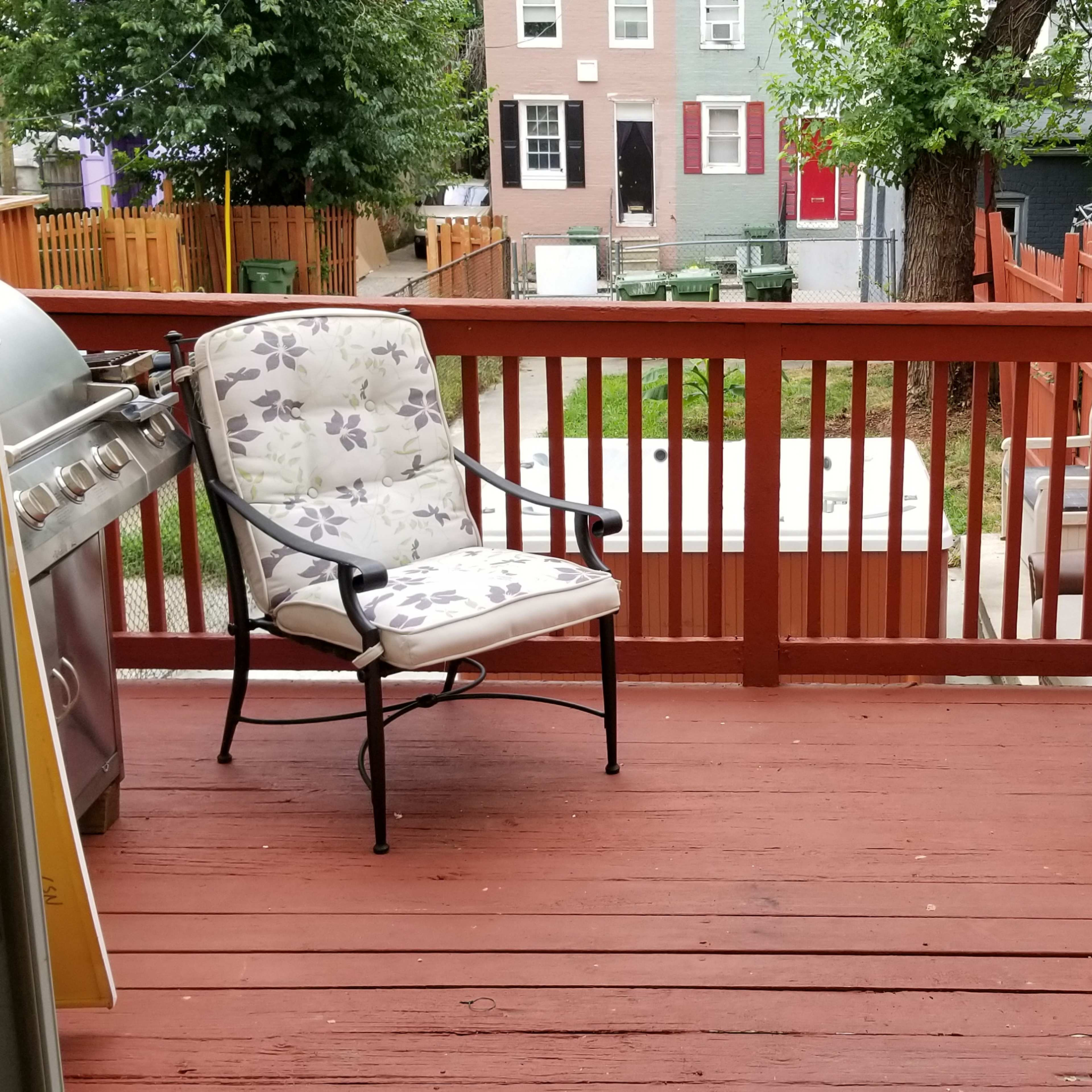 A view from an open door onto a wooden deck with two chairs and a barbecue grill, surrounded by greenery and adjacent buildings.