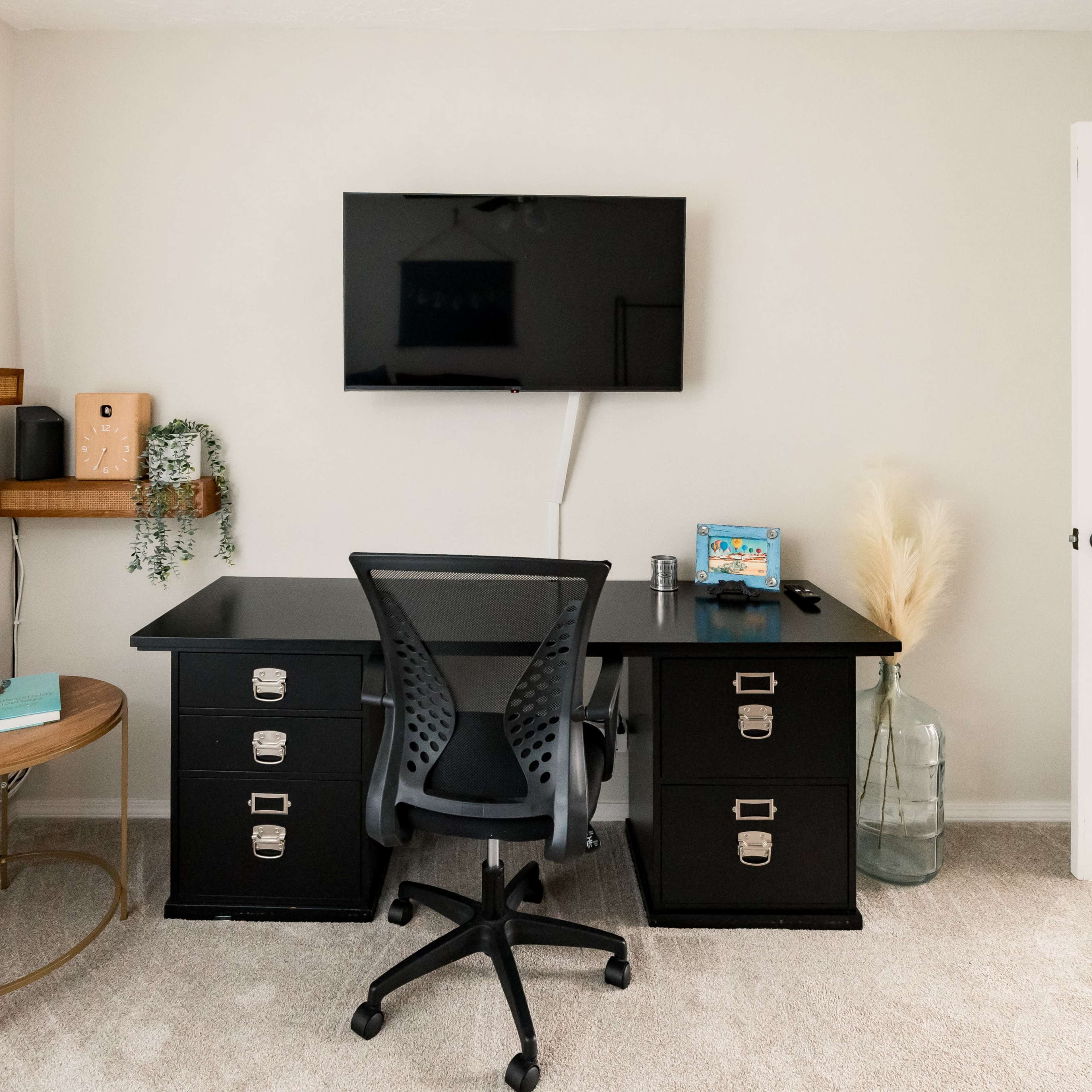 A minimalist office space features a black desk with storage drawers, a chair, a mounted TV on the wall, and a small round table beside it.