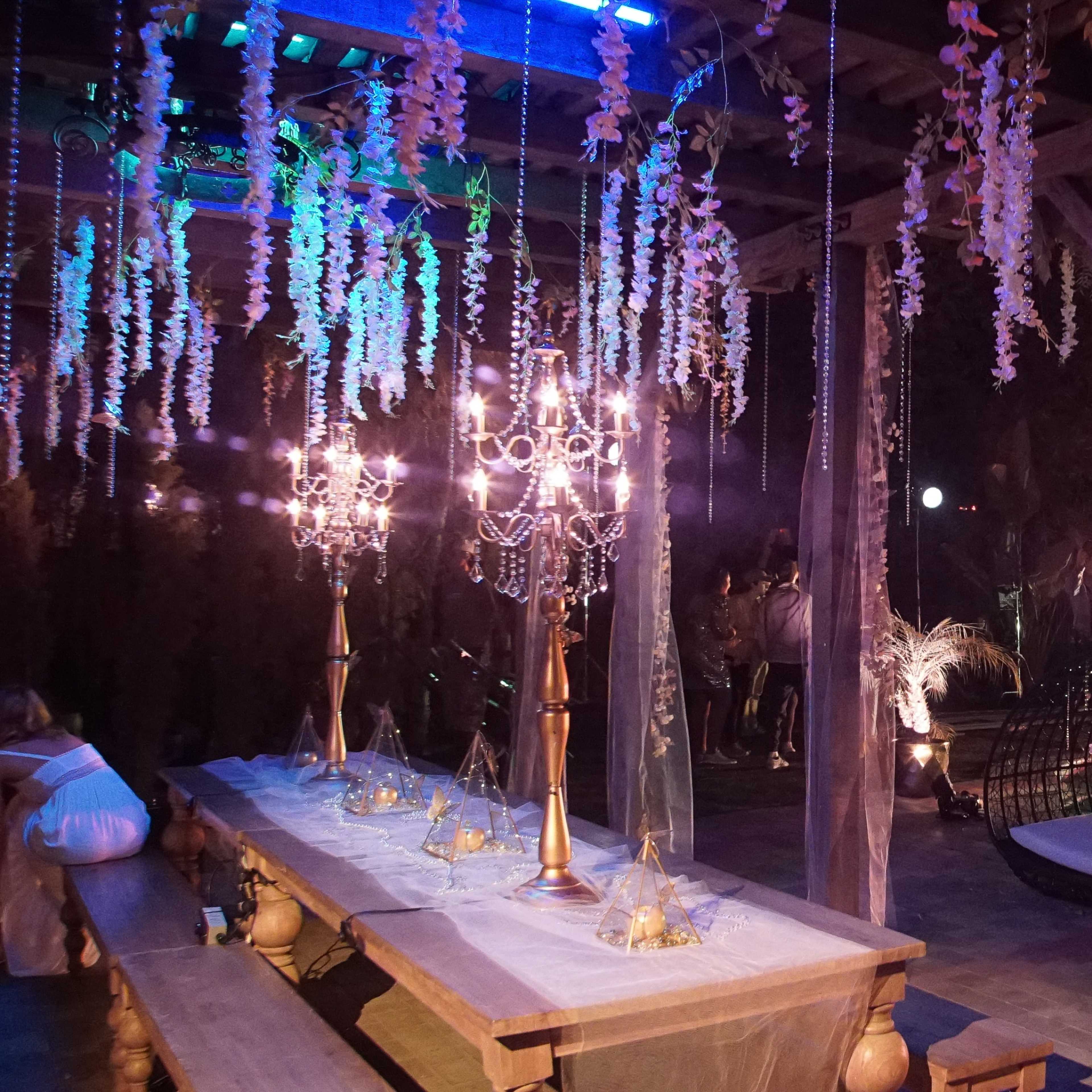 A beautifully decorated outdoor space featuring a long dining table, elegant chandeliers, and a pool surrounded by tropical foliage.