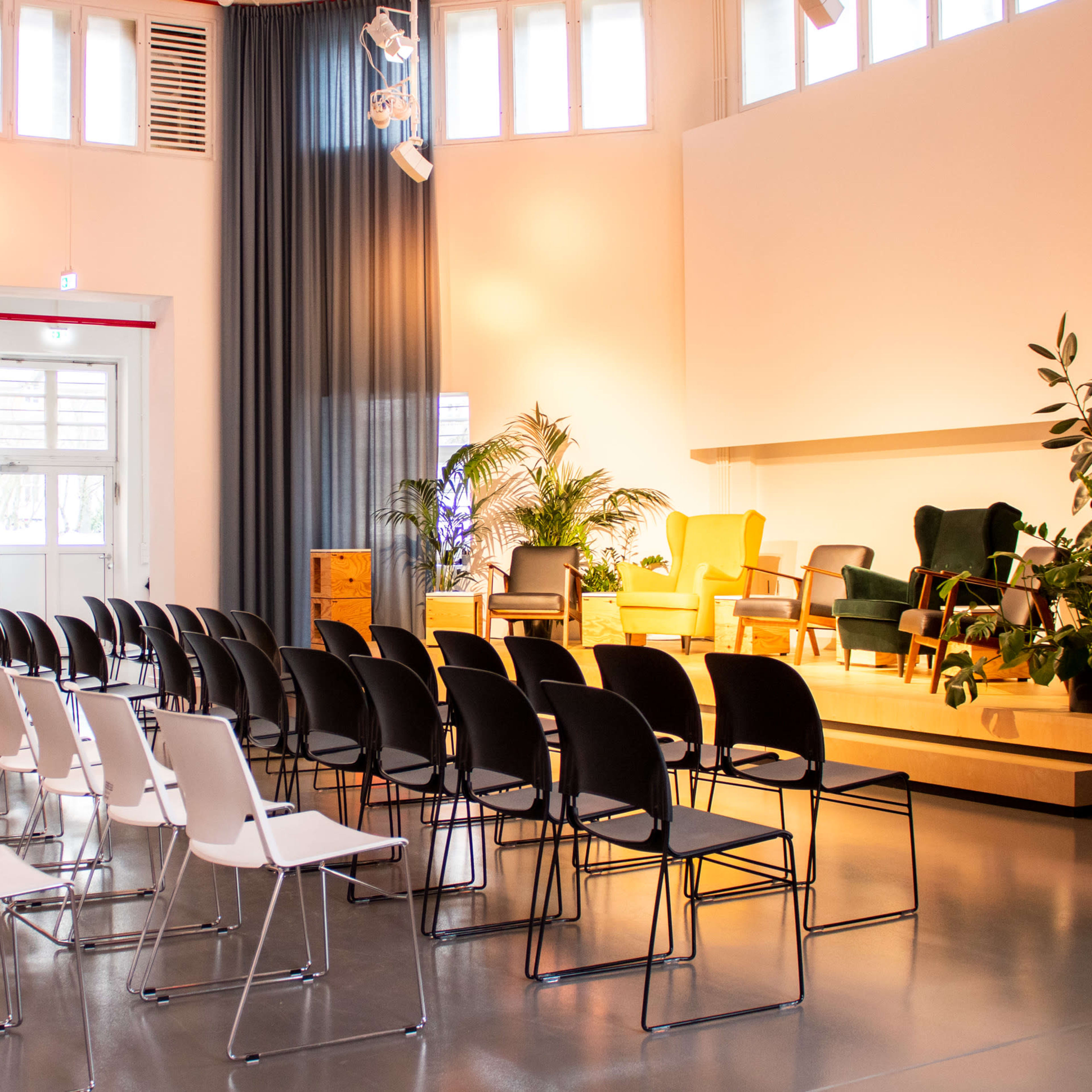 A seminar room with several rows of empty chairs facing a stage set with two armchairs and greenery.