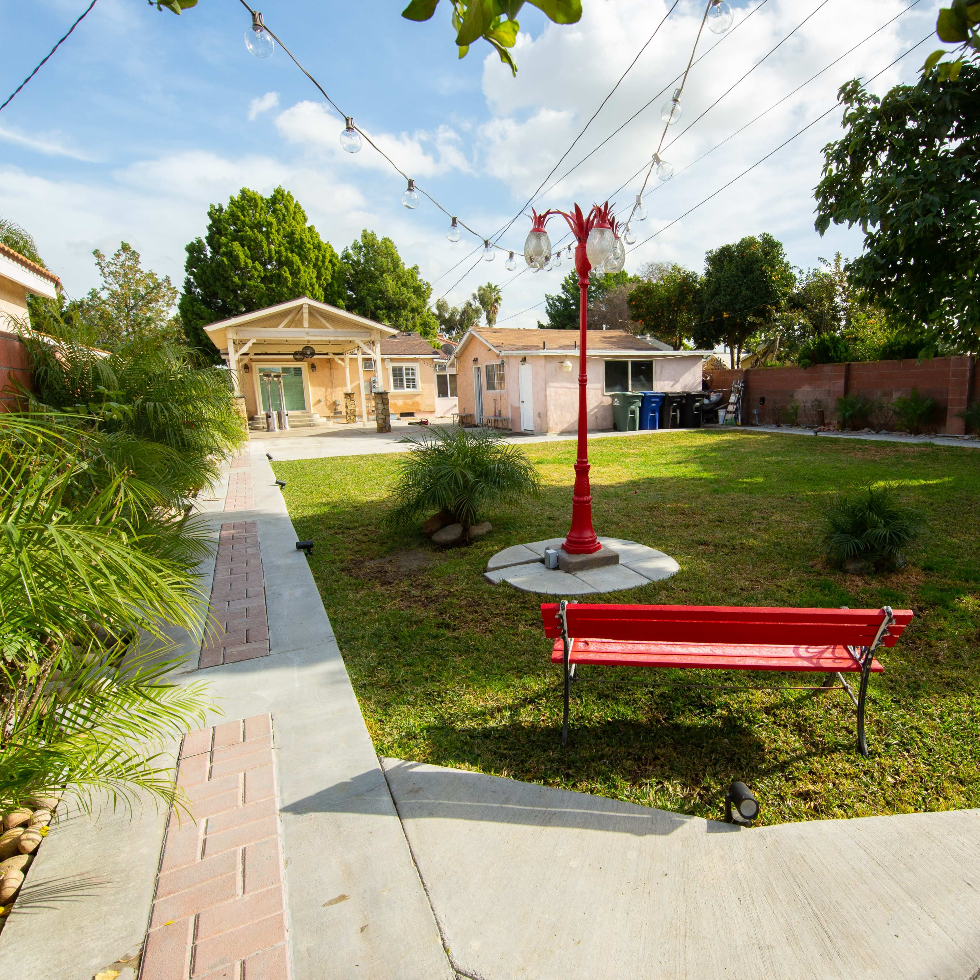 A red bench sits near a lamp post in a grassy yard surrounded by landscaped pathways and small trees.