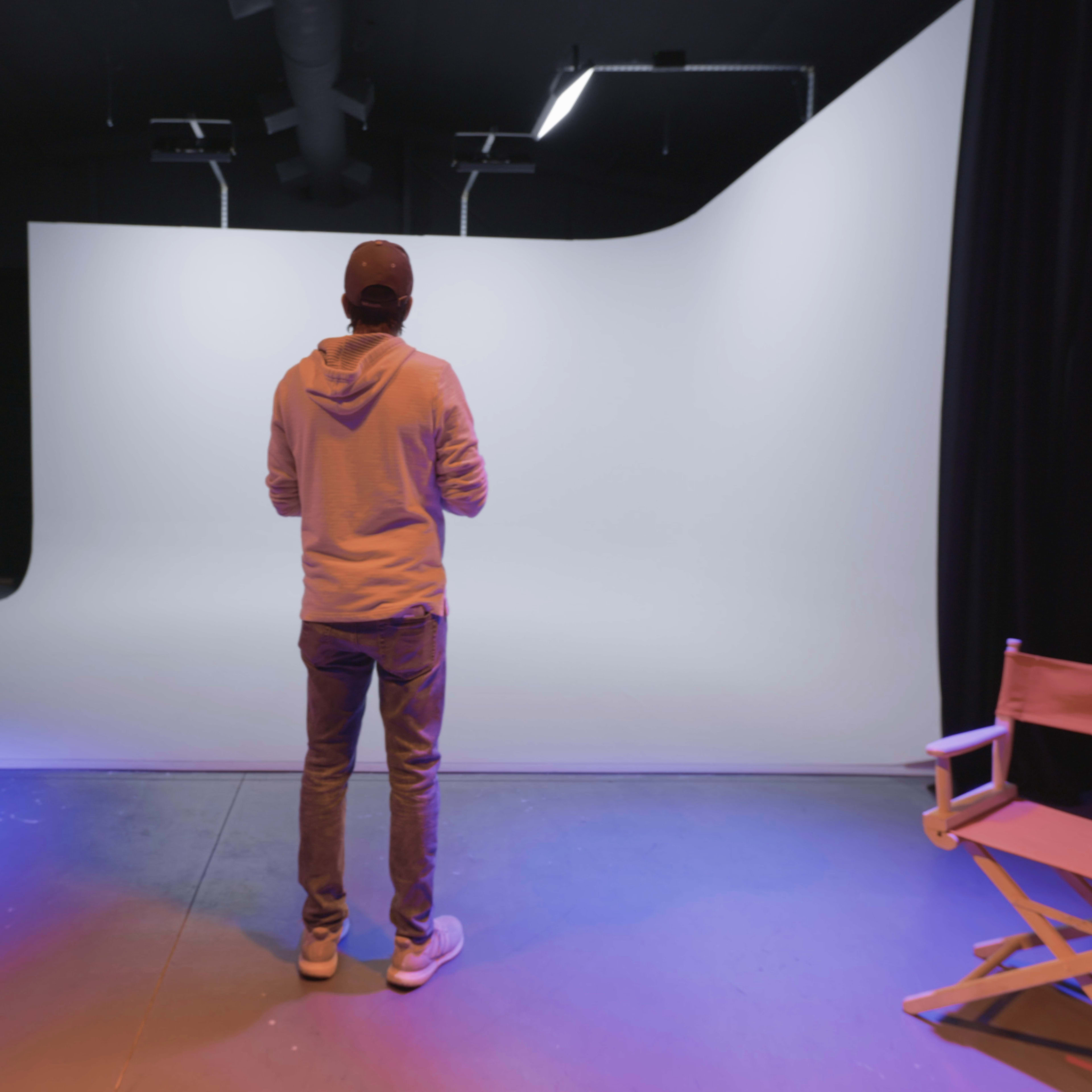 A person stands in front of a blank white backdrop in a studio, with a director's chair visible to the side.
