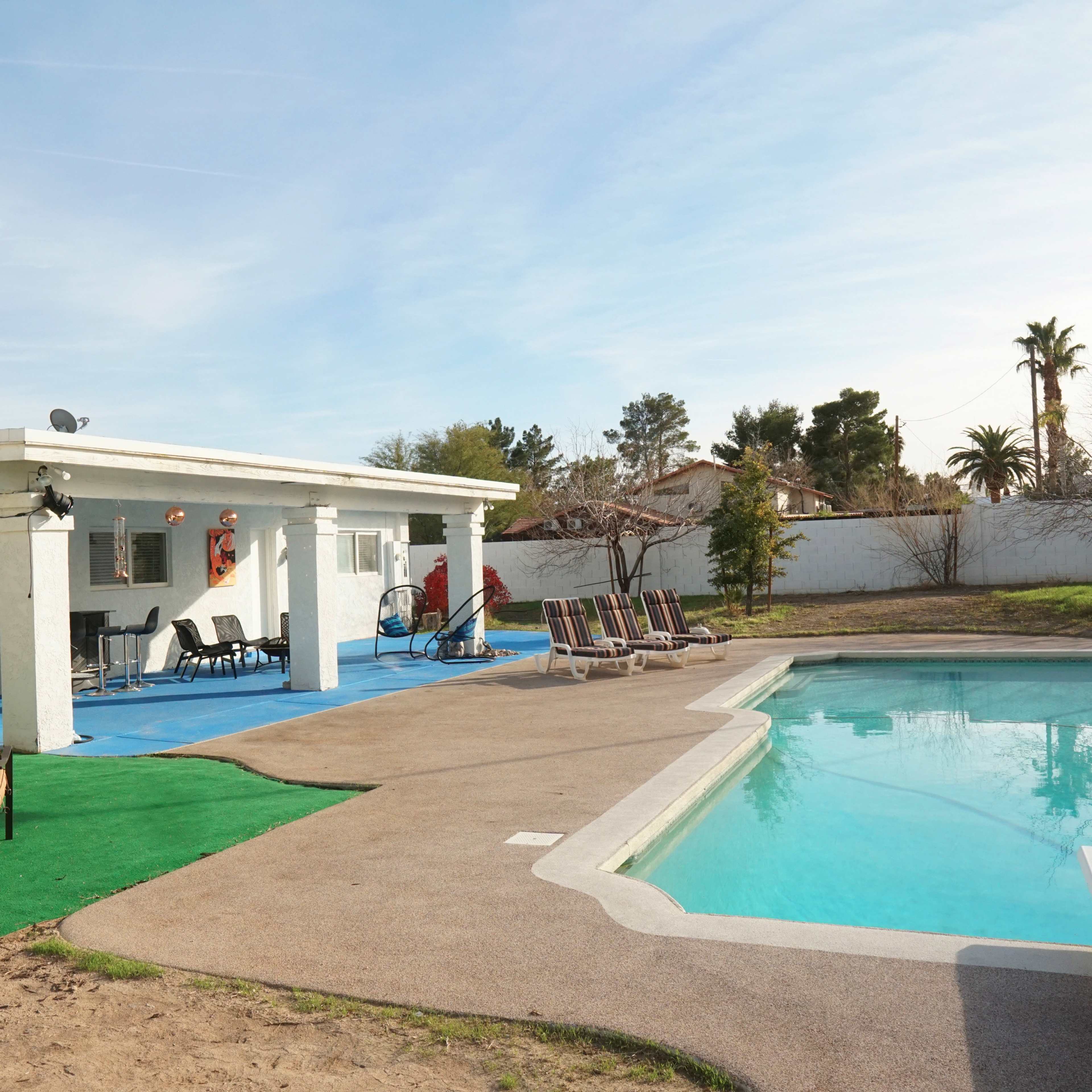 A patio area with outdoor seating and a pool is located next to a single-story house surrounded by a grassy lawn and palm trees.