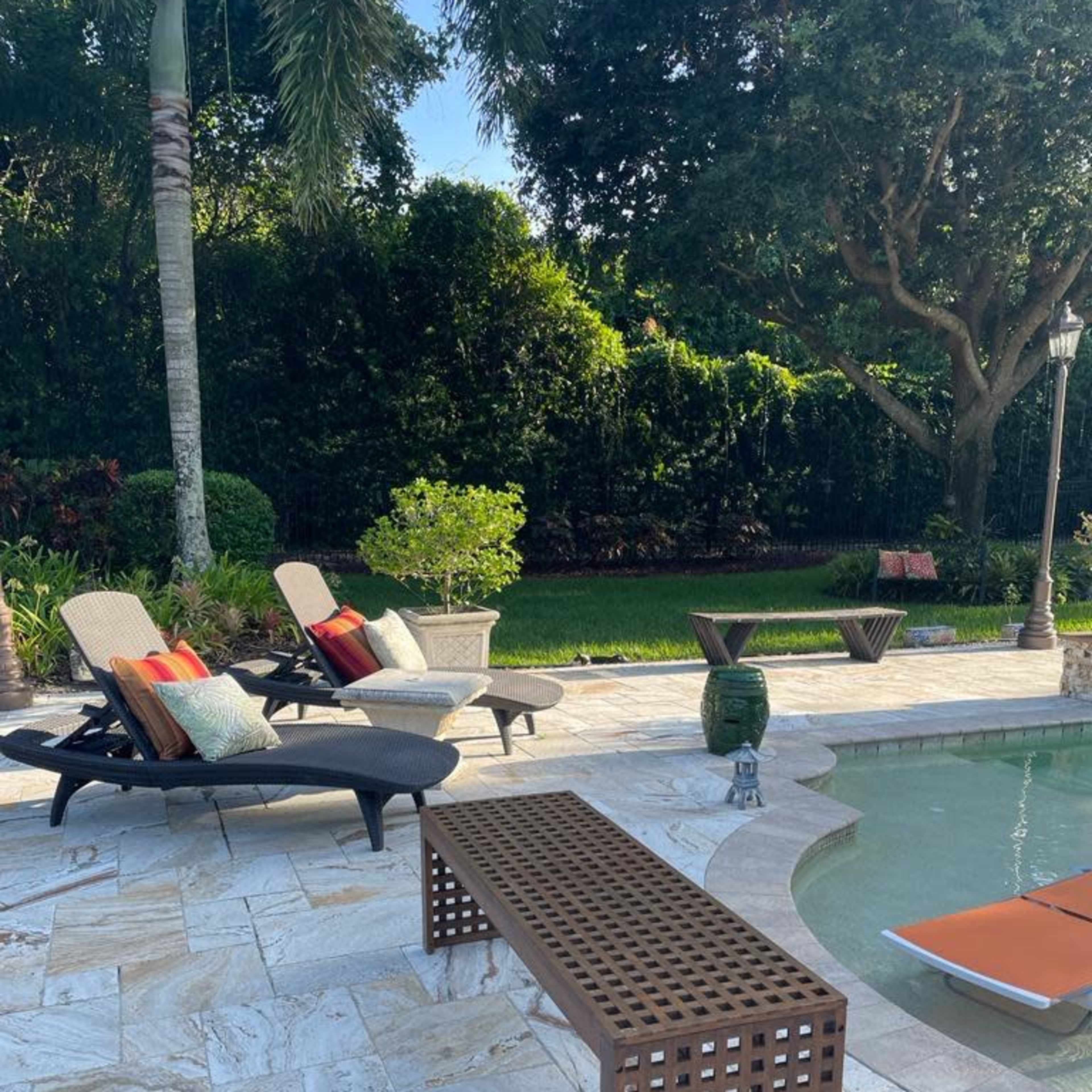 The scene features a poolside area with two lounge chairs, a decorative bench, and a lush green backdrop.