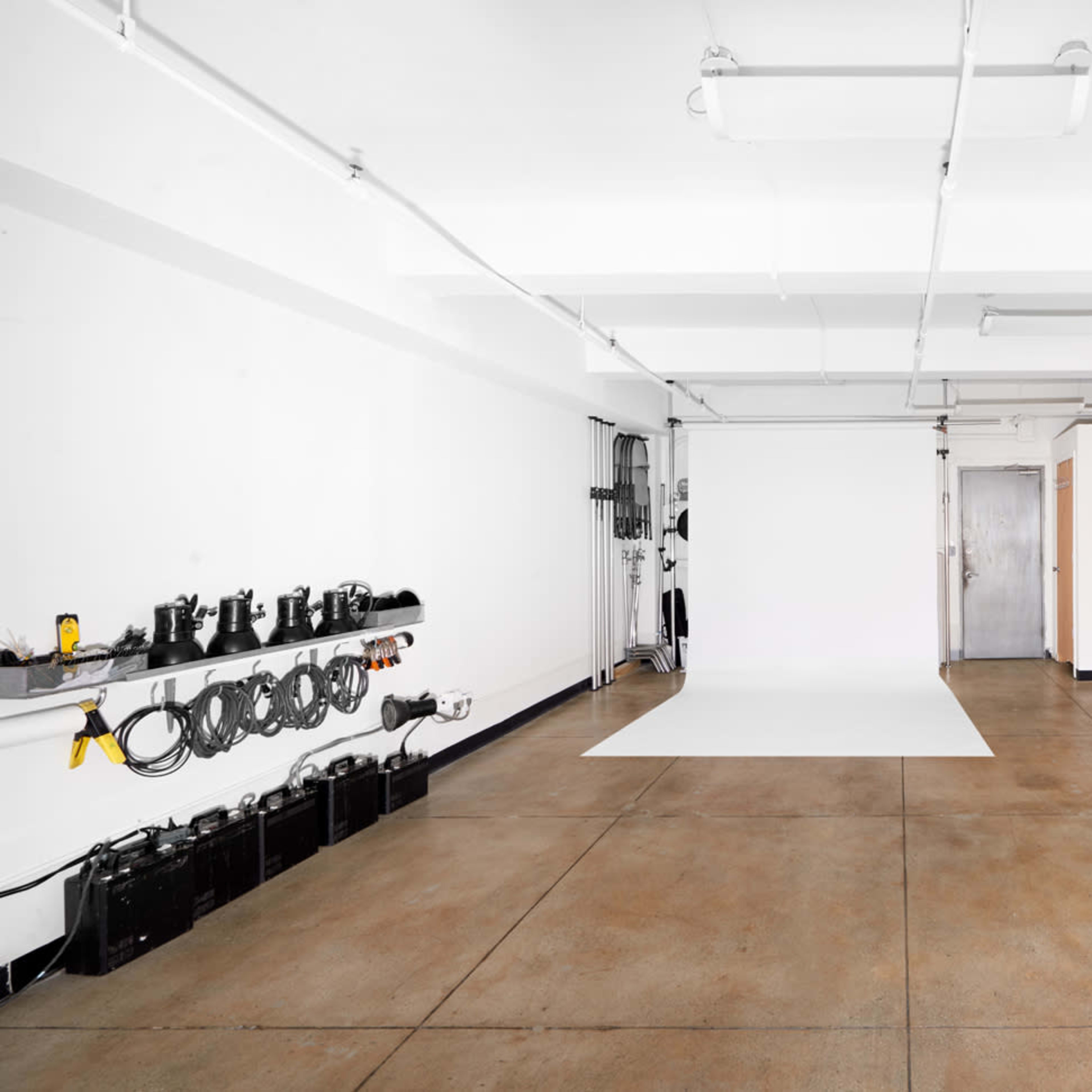 A spacious, empty studio with a white backdrop and industrial flooring, equipped with lighting and equipment mounted along the wall.