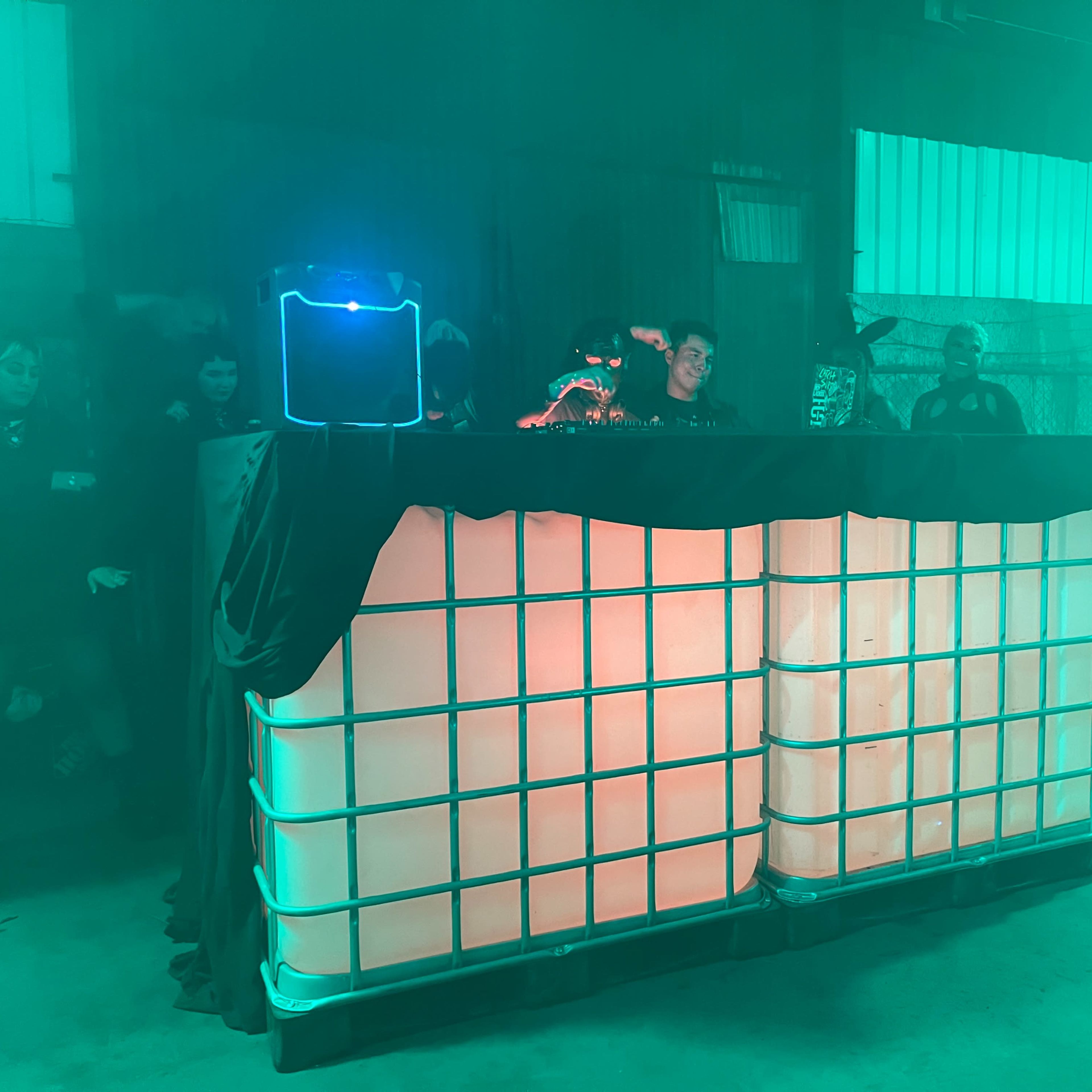 A DJ booth made of stacked white containers is illuminated with green and orange lighting in a warehouse setting.