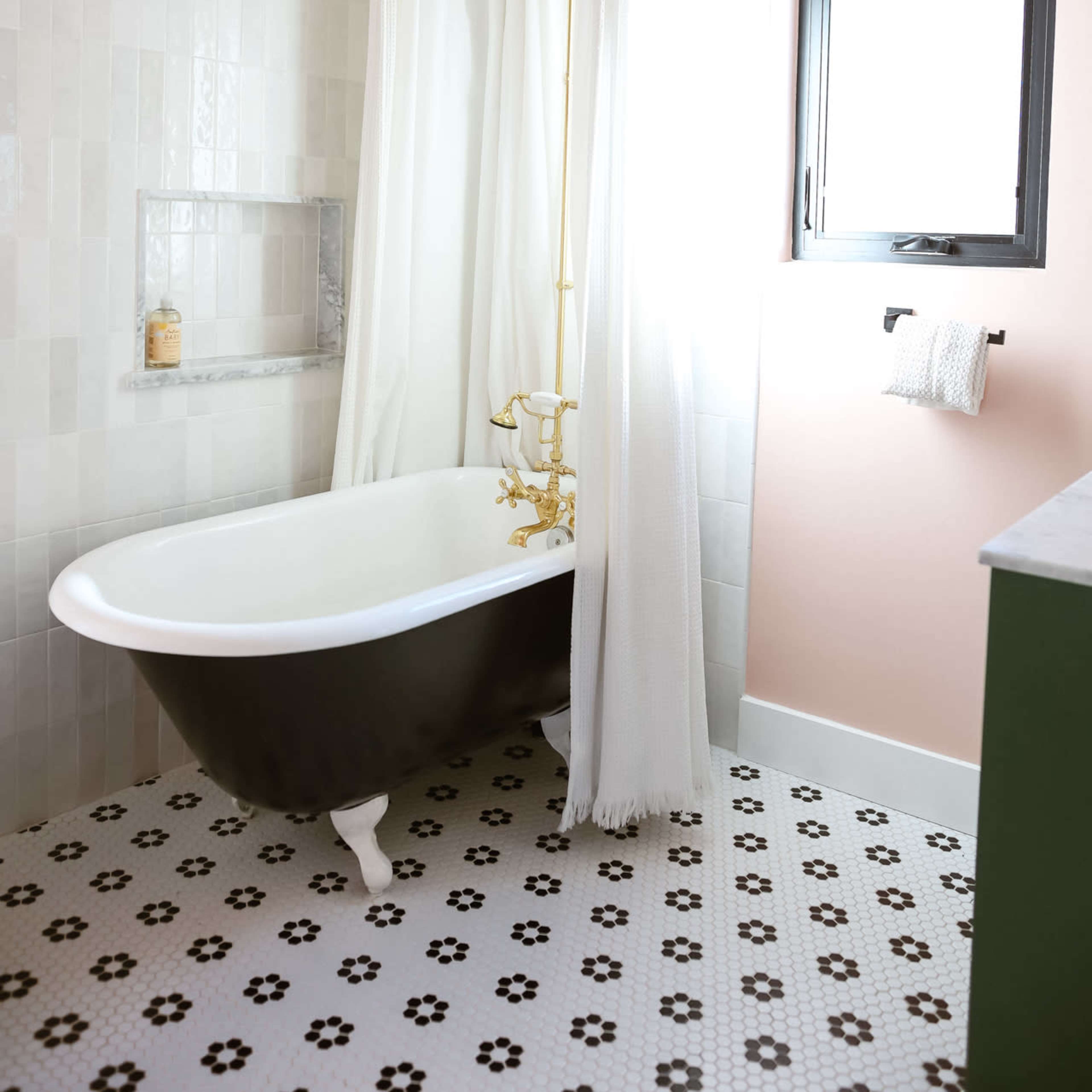 A bathroom with a freestanding bathtub, a gold shower rod, and a light-colored tile wall alongside a window with a black frame.