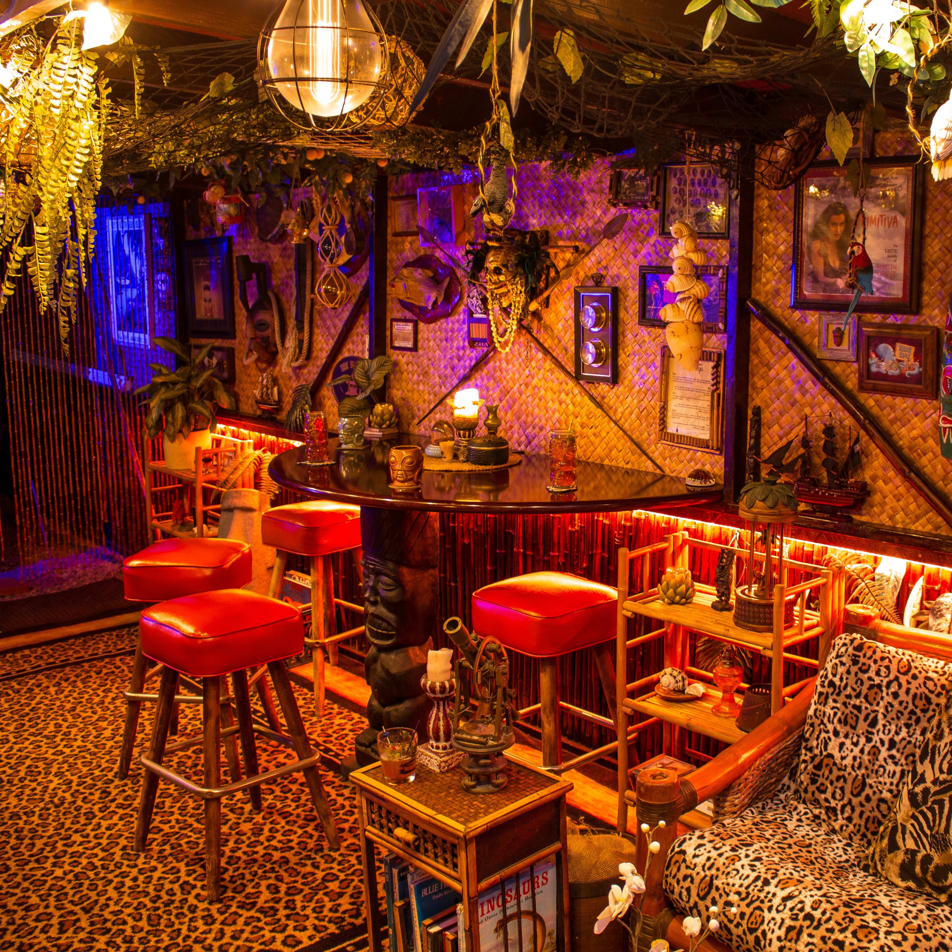 A decorated indoor lounge with a bar, surrounded by tropical plants, animal prints, and various framed artworks.