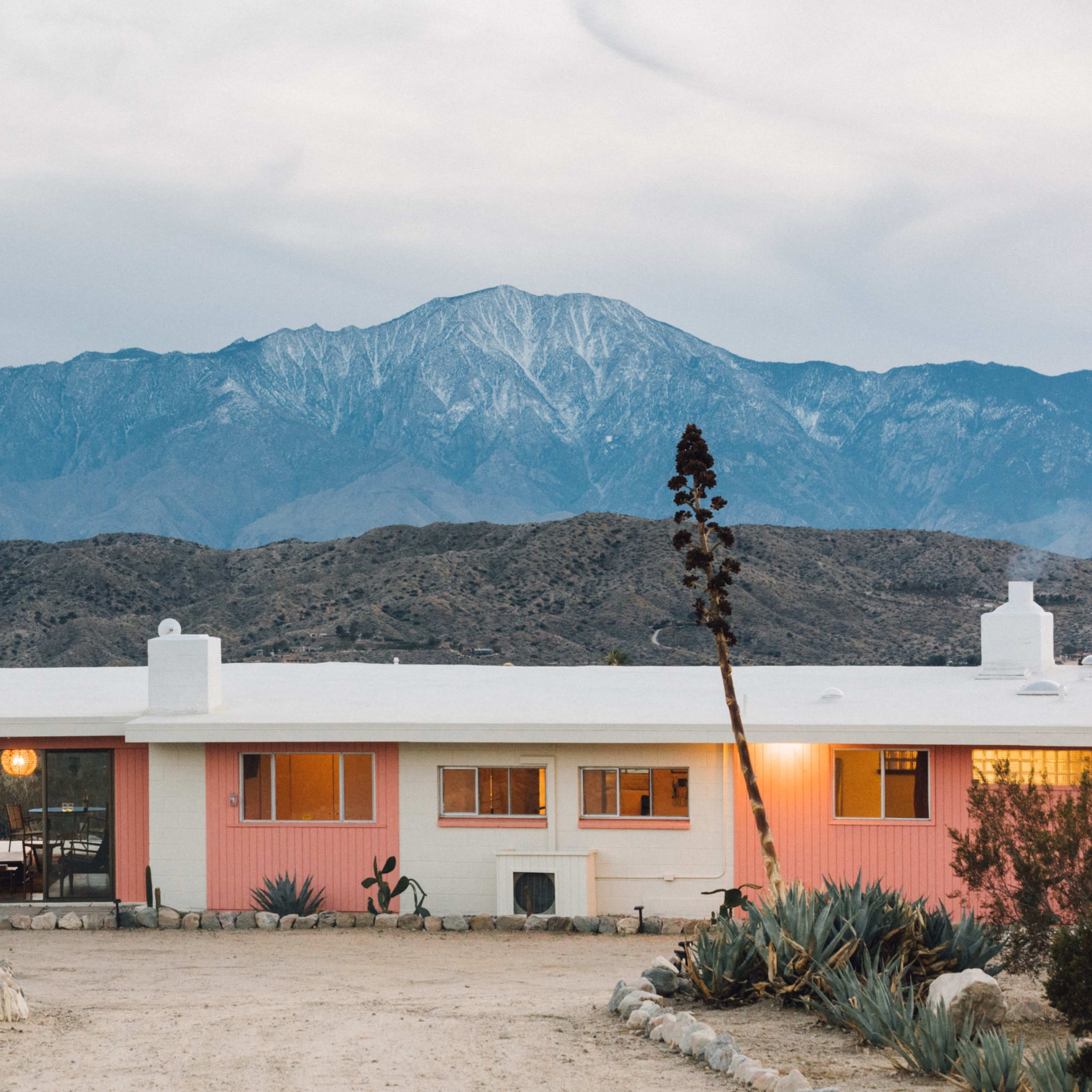 A pink mid-century modern house stands in front of a mountainous landscape with snow-capped peaks.