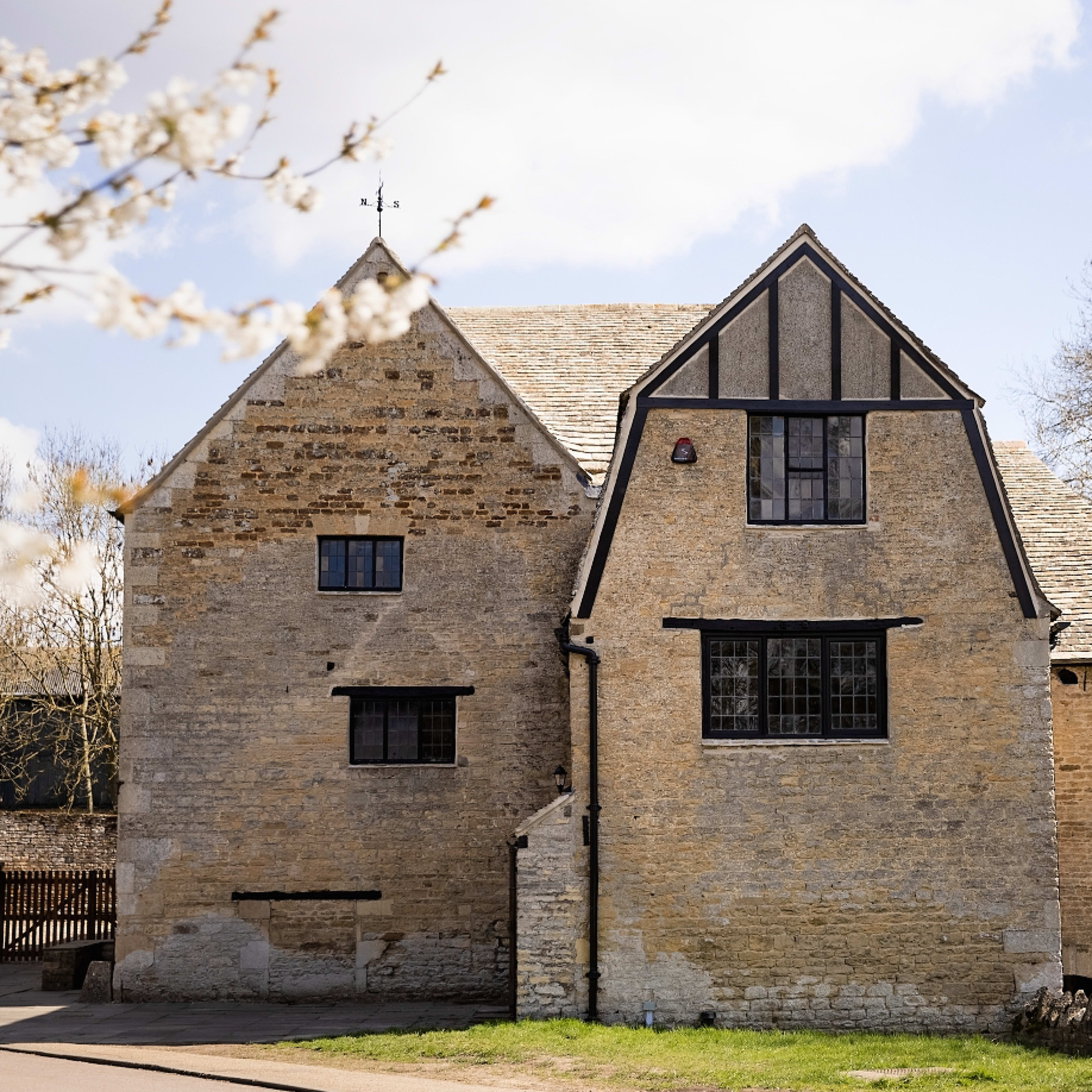 A stone and timber-framed house stands beside a road, surrounded by trees and blooming plants.