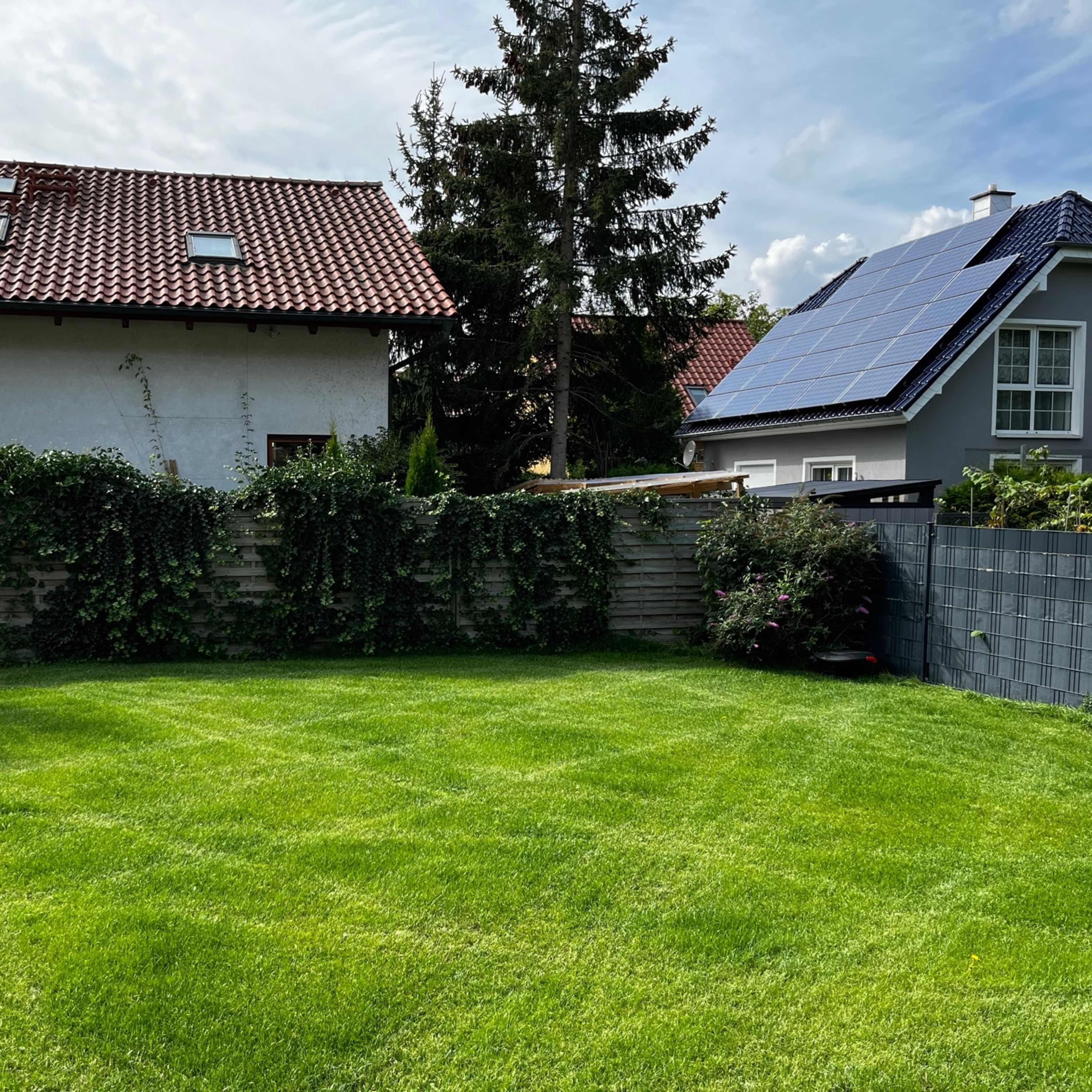 A well-maintained green lawn surrounded by fenced garden areas and two houses with solar panels and a swing set.