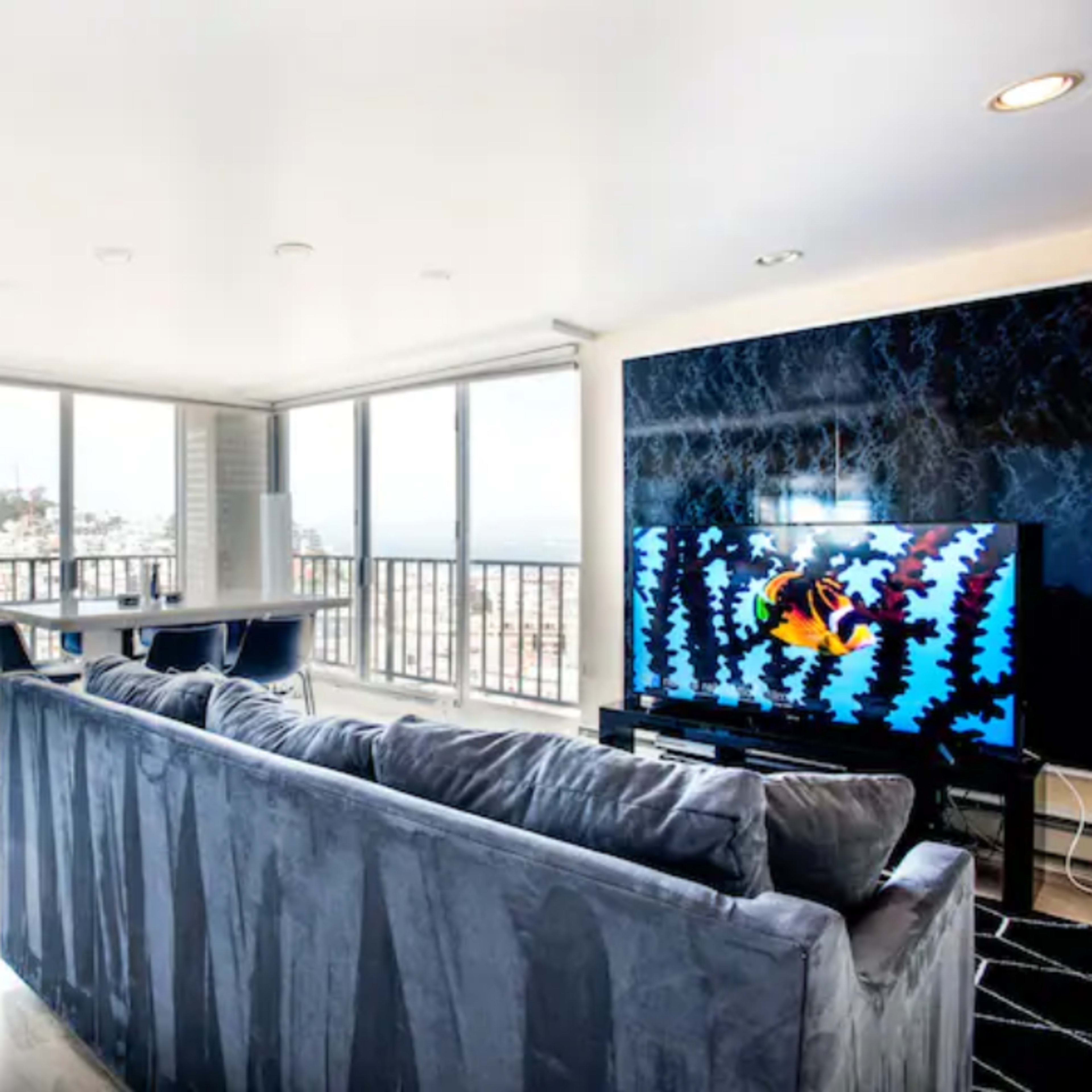 A modern living room with a large couch facing a wall-mounted television and large windows offering a view of the city.