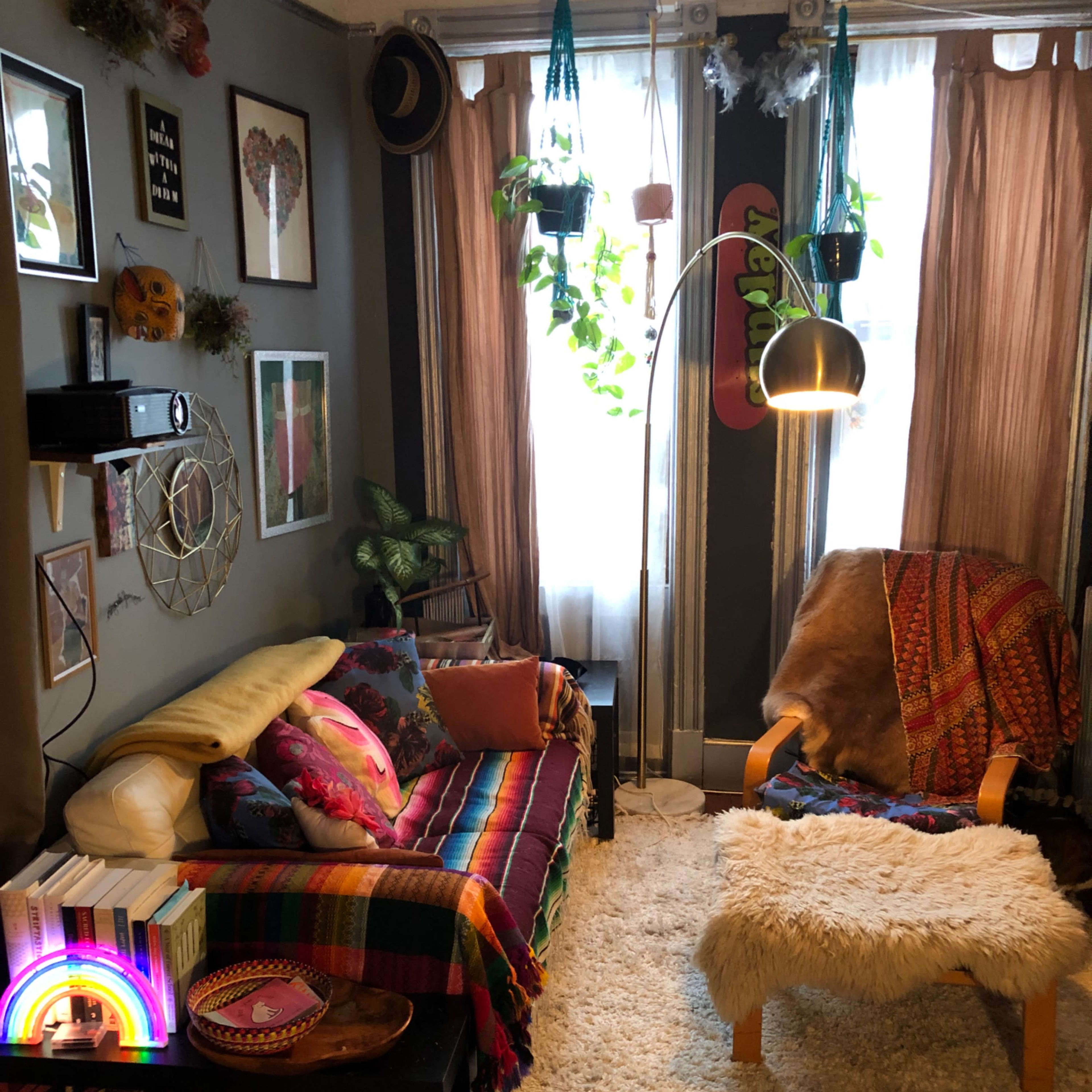 A cozy living room with a colorful sofa, a wooden chair, and various decorative plants hanging near a large window.