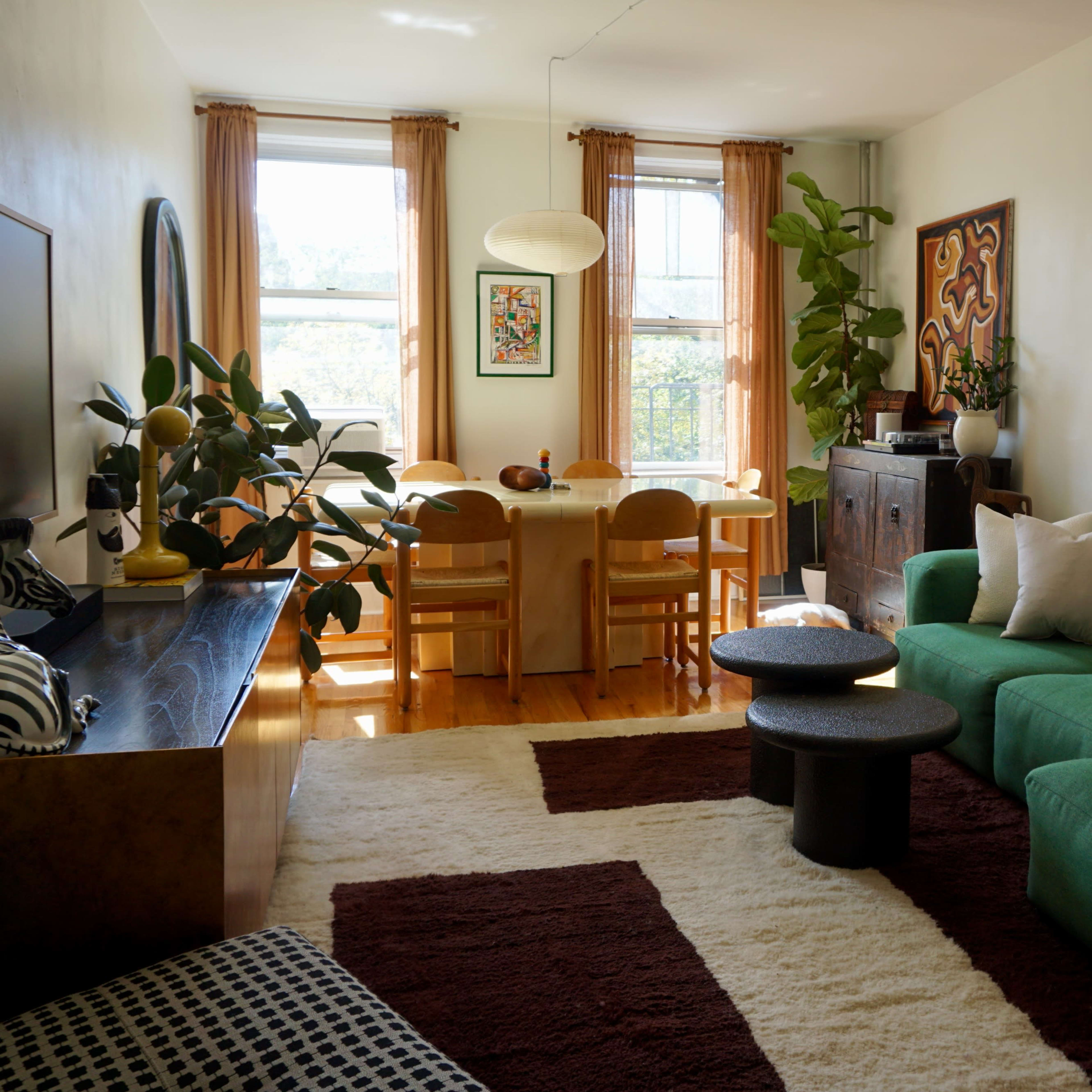 The room features a living area with a green sectional sofa, a black and white coffee table, and a dining table with wooden chairs, accompanied by large windows and plants.