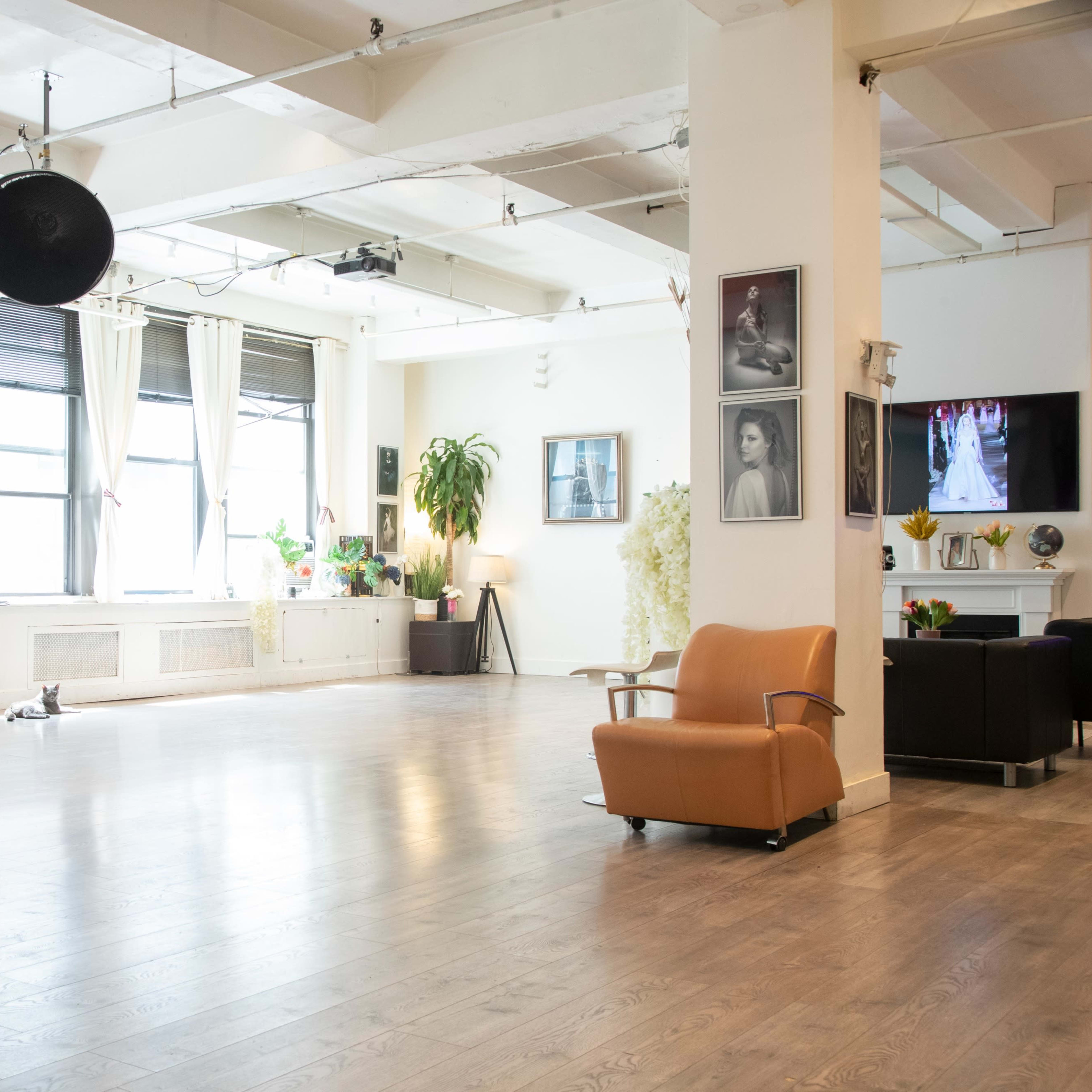 A well-lit studio space with large windows, wooden flooring, and minimal furniture including an orange chair.