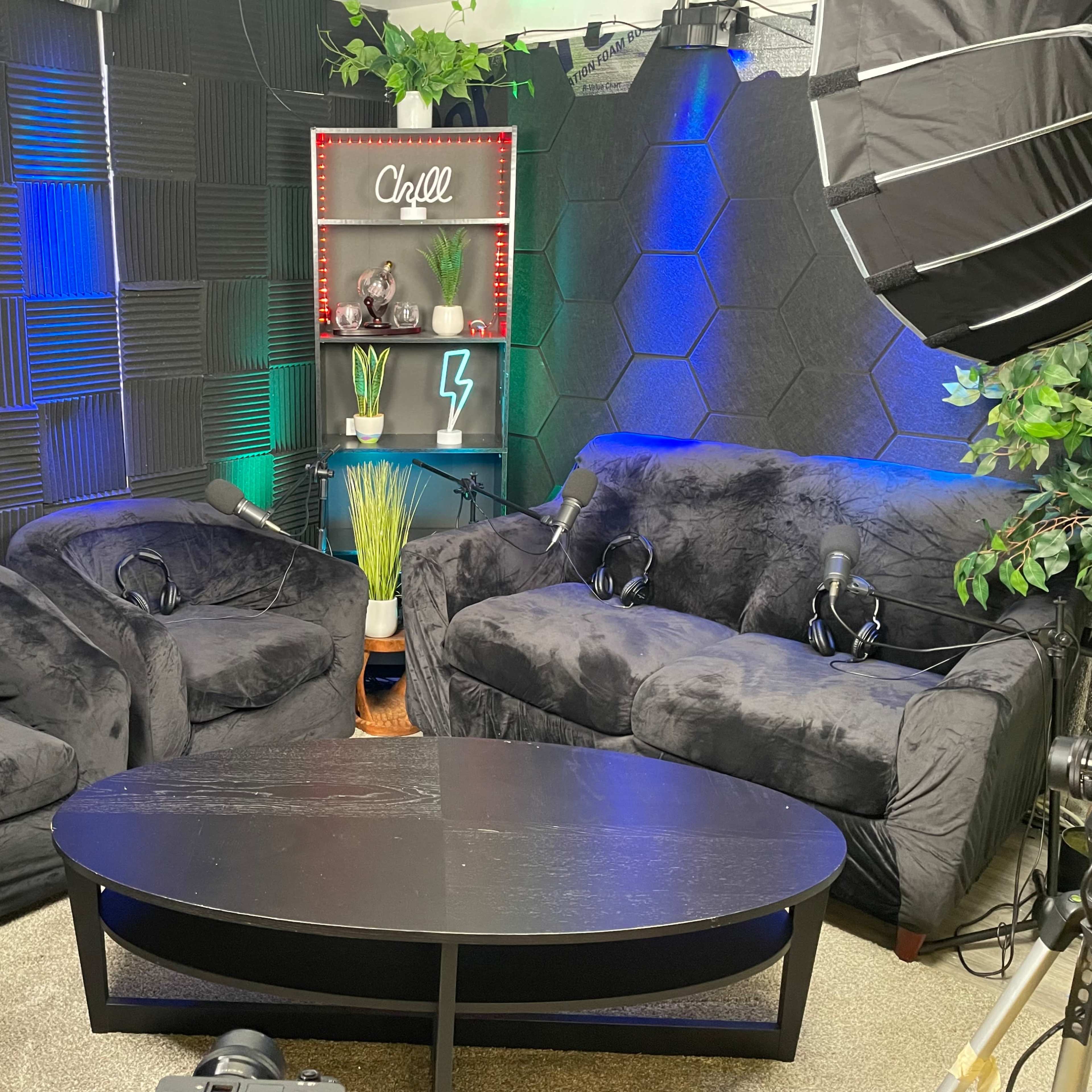 A cozy room setup with two black couches, a dark coffee table, and a backdrop featuring decorative plants and lighting, intended for a podcast or interview.