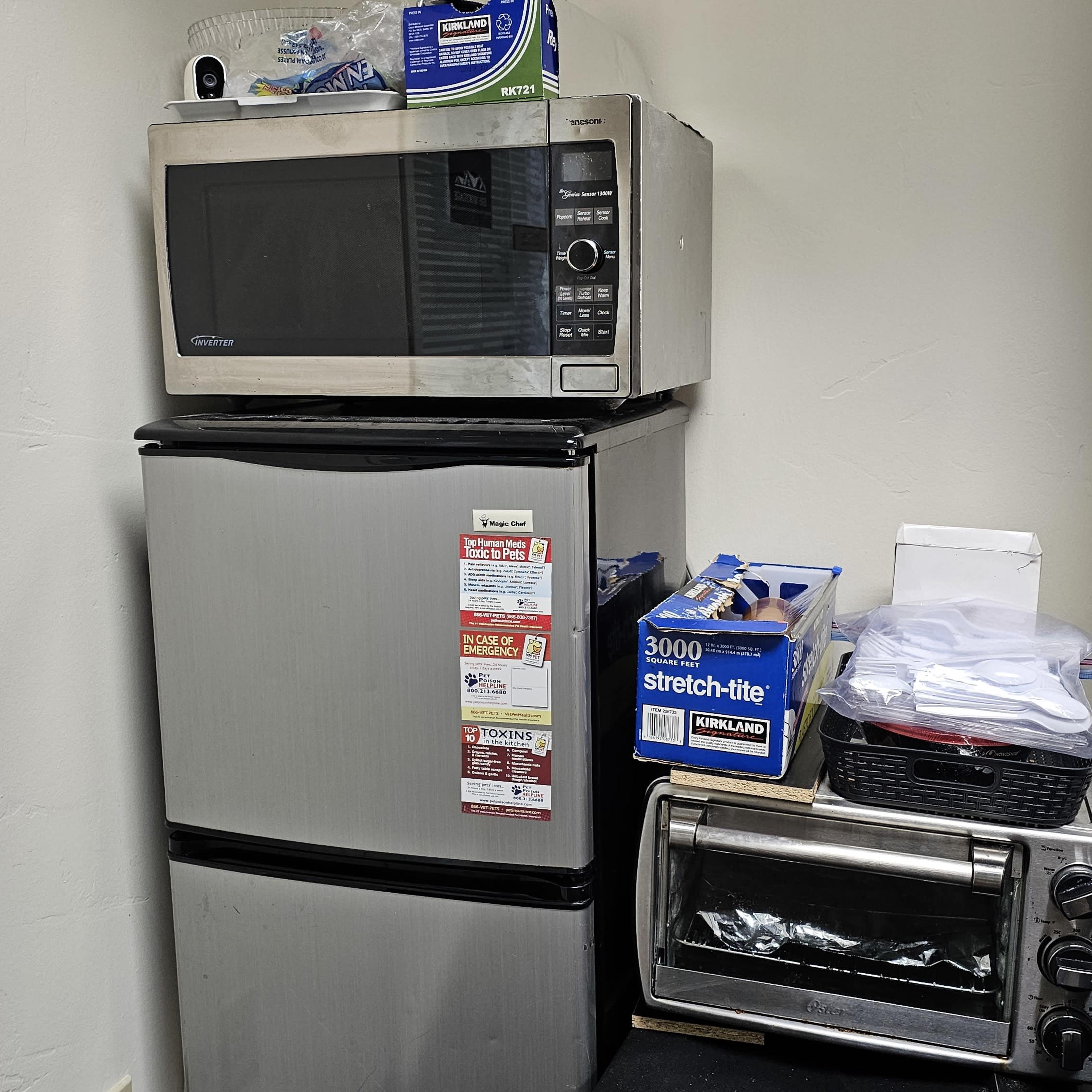 A microwave is placed on top of a compact refrigerator, with kitchen items stacked nearby, including a toaster oven and some bags.