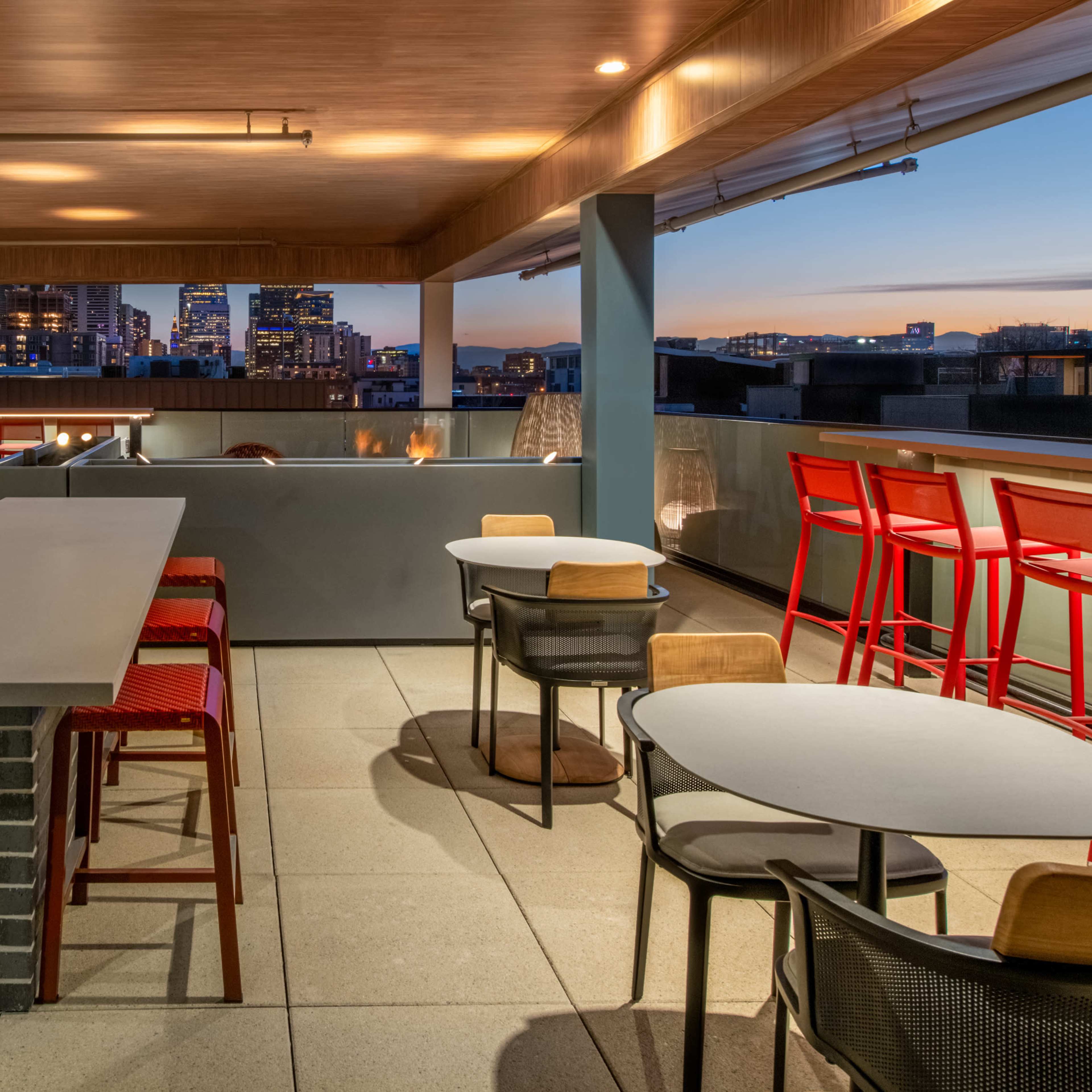 A modern rooftop terrace featuring a grill, bar seating, and tables with city skyline views at dusk.