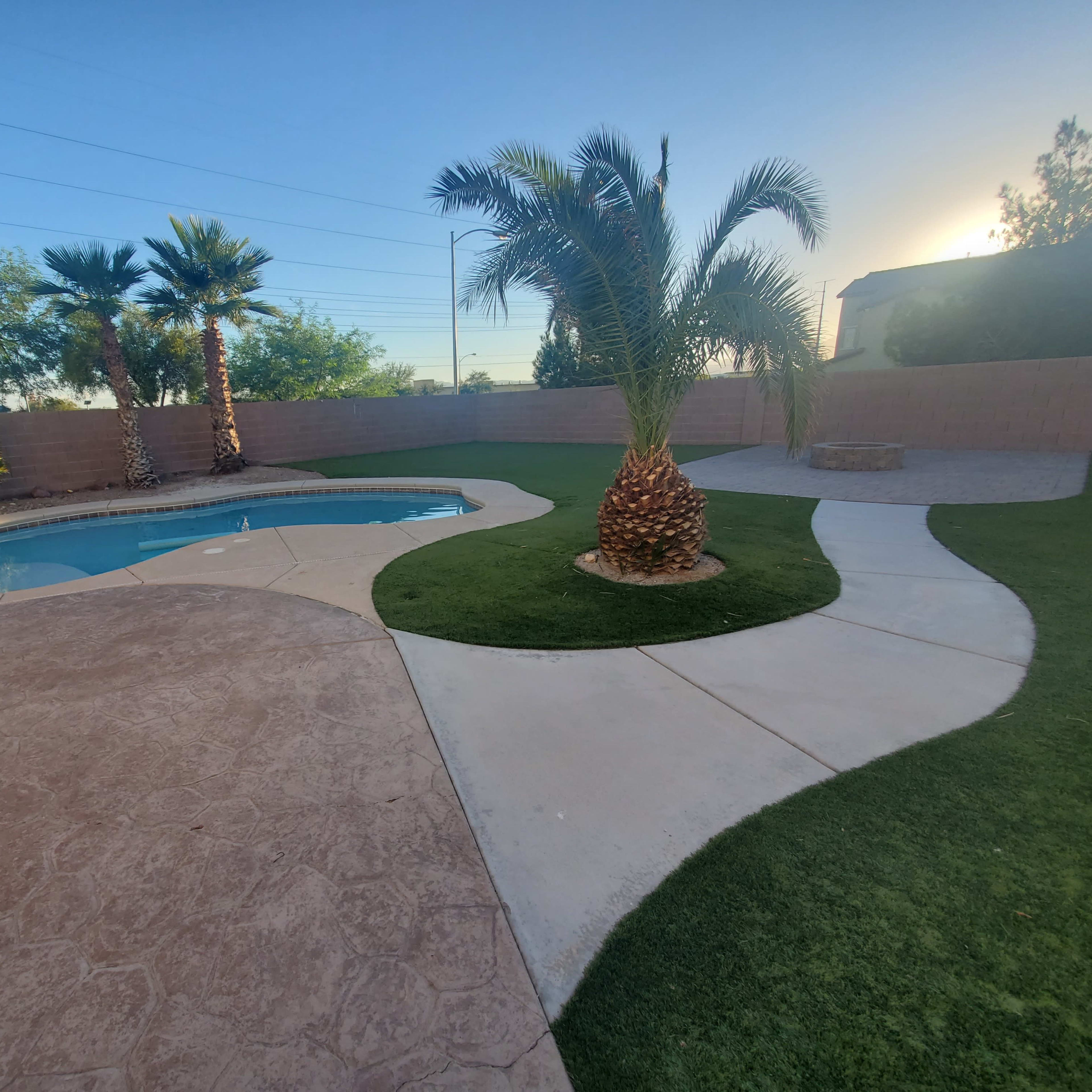 A backyard with a swimming pool surrounded by landscaped grass, palm trees, and a stone fire pit.