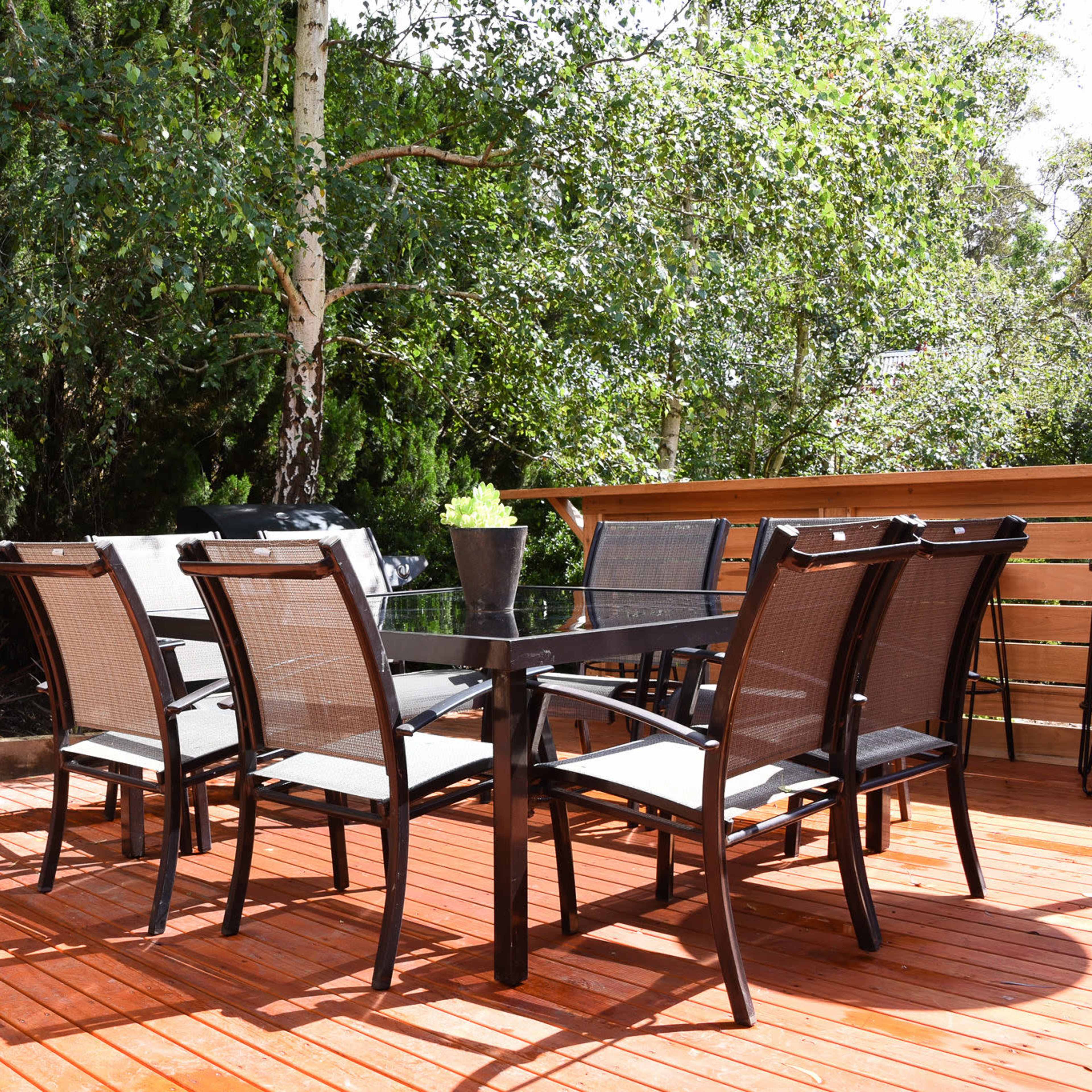 A wooden deck features a table surrounded by six black mesh chairs, set in a lush green garden.