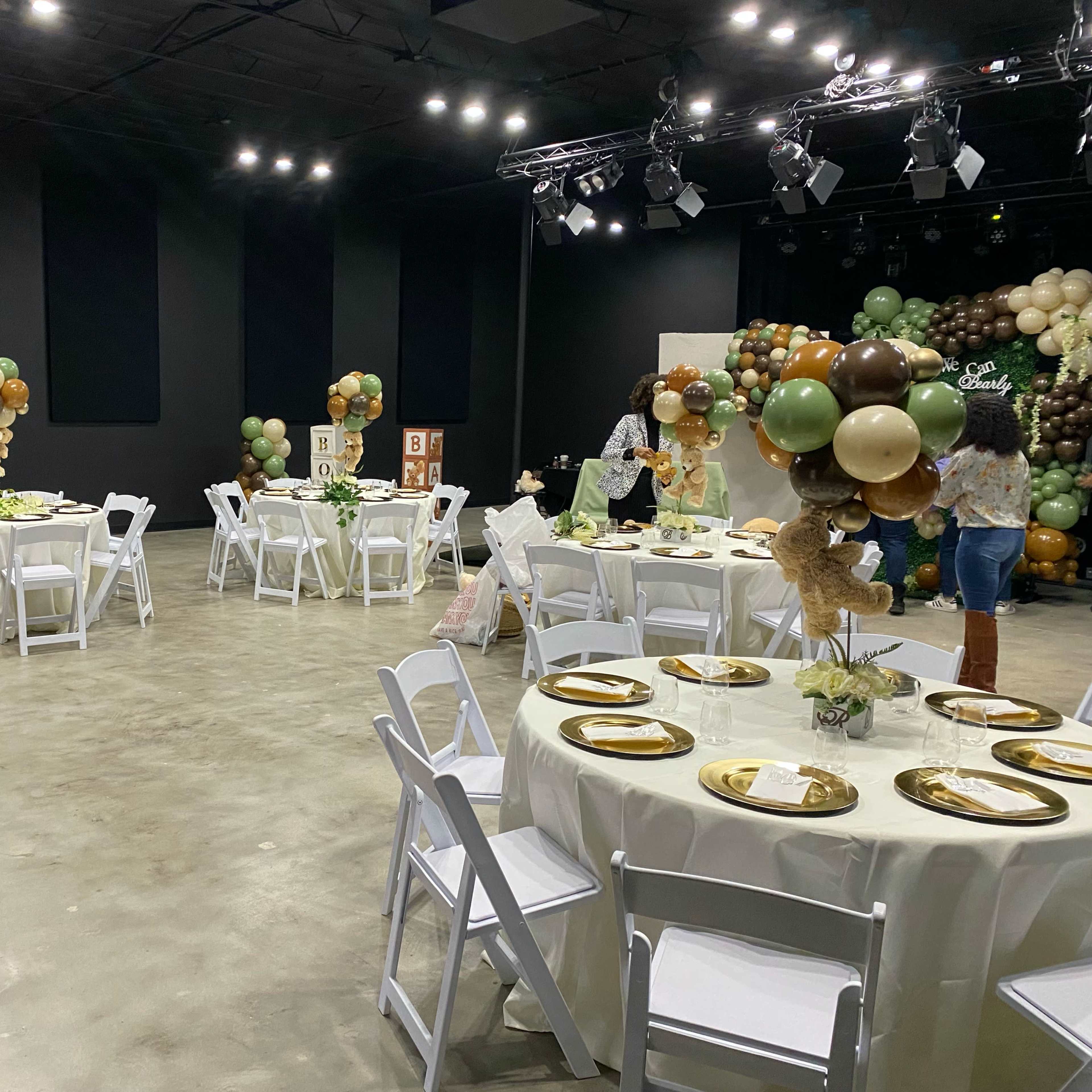 A decorated event space features several round tables set with gold plates and flowers, surrounded by large balloon arrangements.