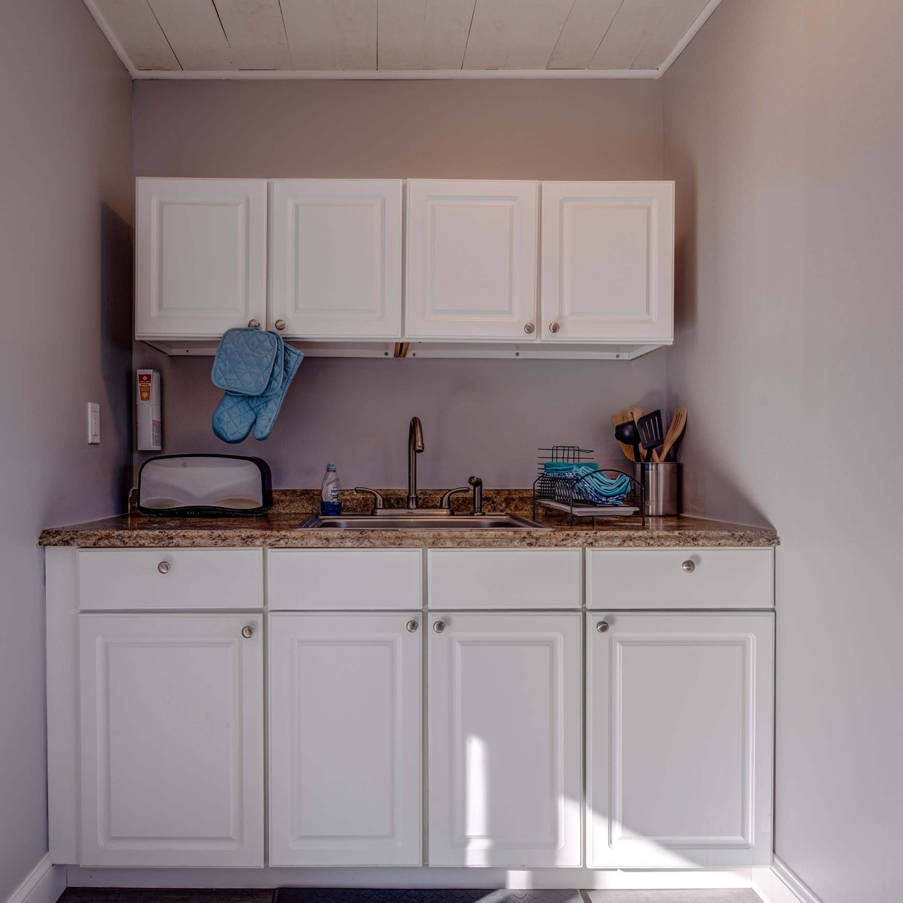 A small kitchen area features white cabinets above a countertop with a sink, a microwave, and utensils in a metal container.