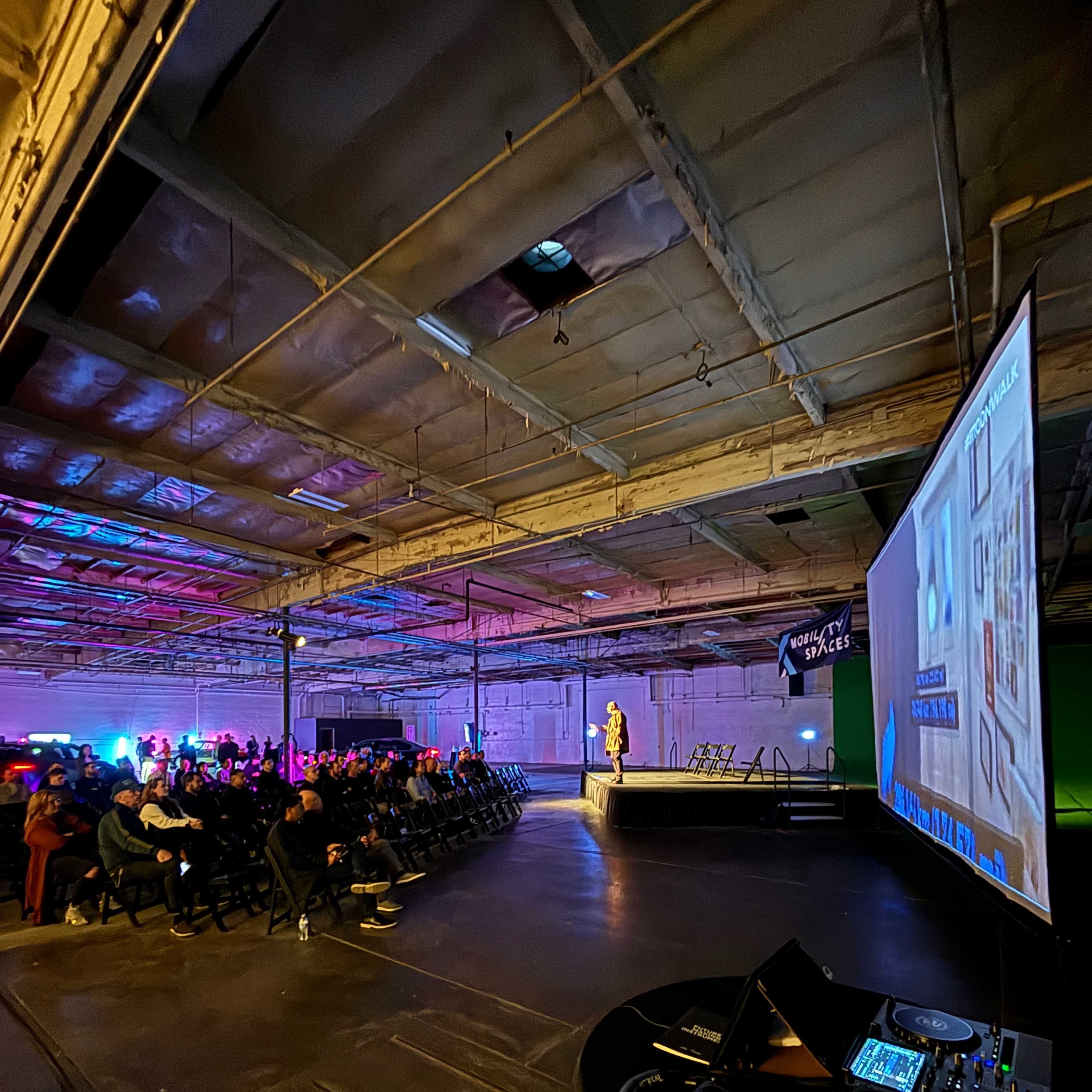 A speaker presents on stage to an audience seated in a dimly lit warehouse, with a large screen displaying visuals beside them.