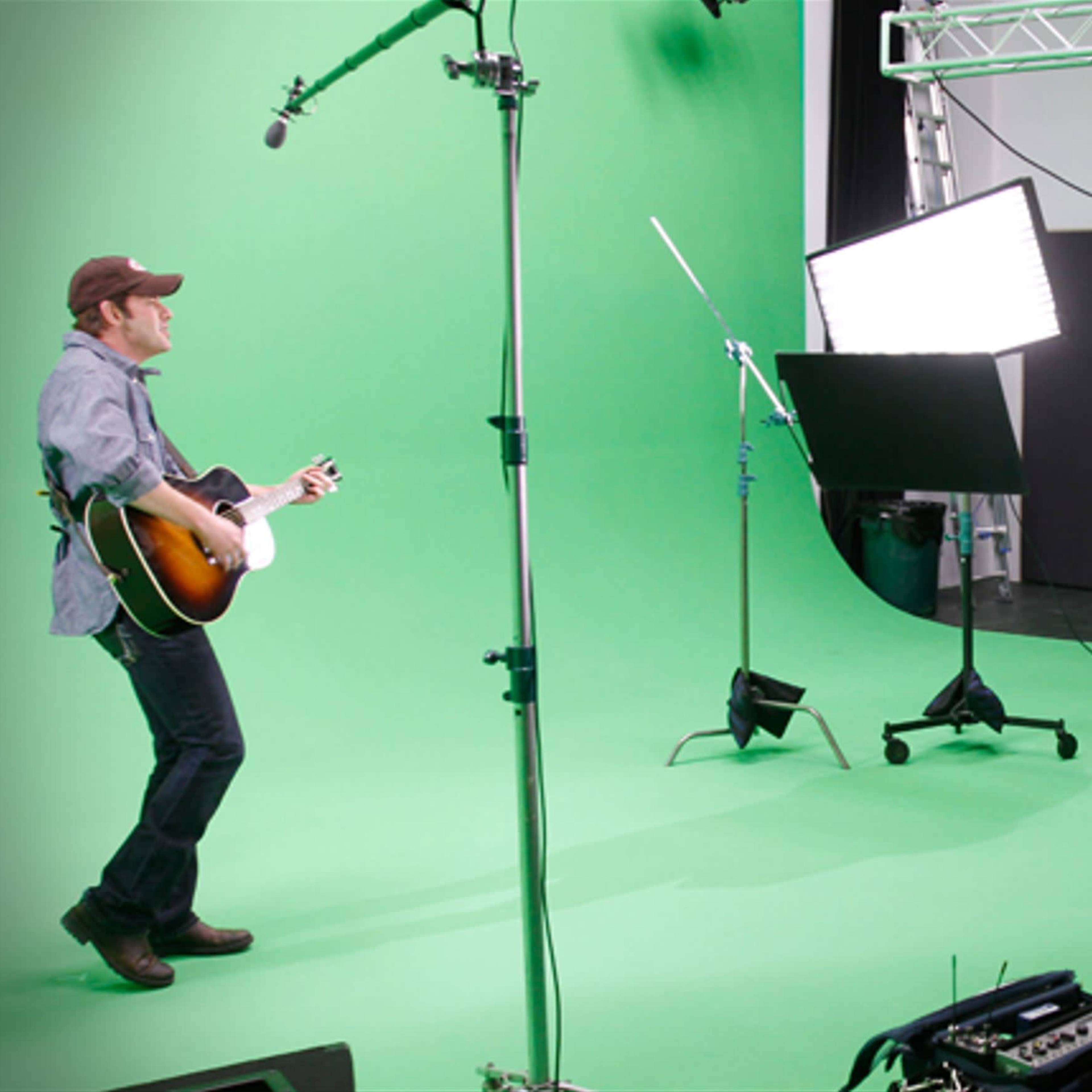 A musician plays an acoustic guitar in front of a green screen while a crew observes from behind a recording setup.