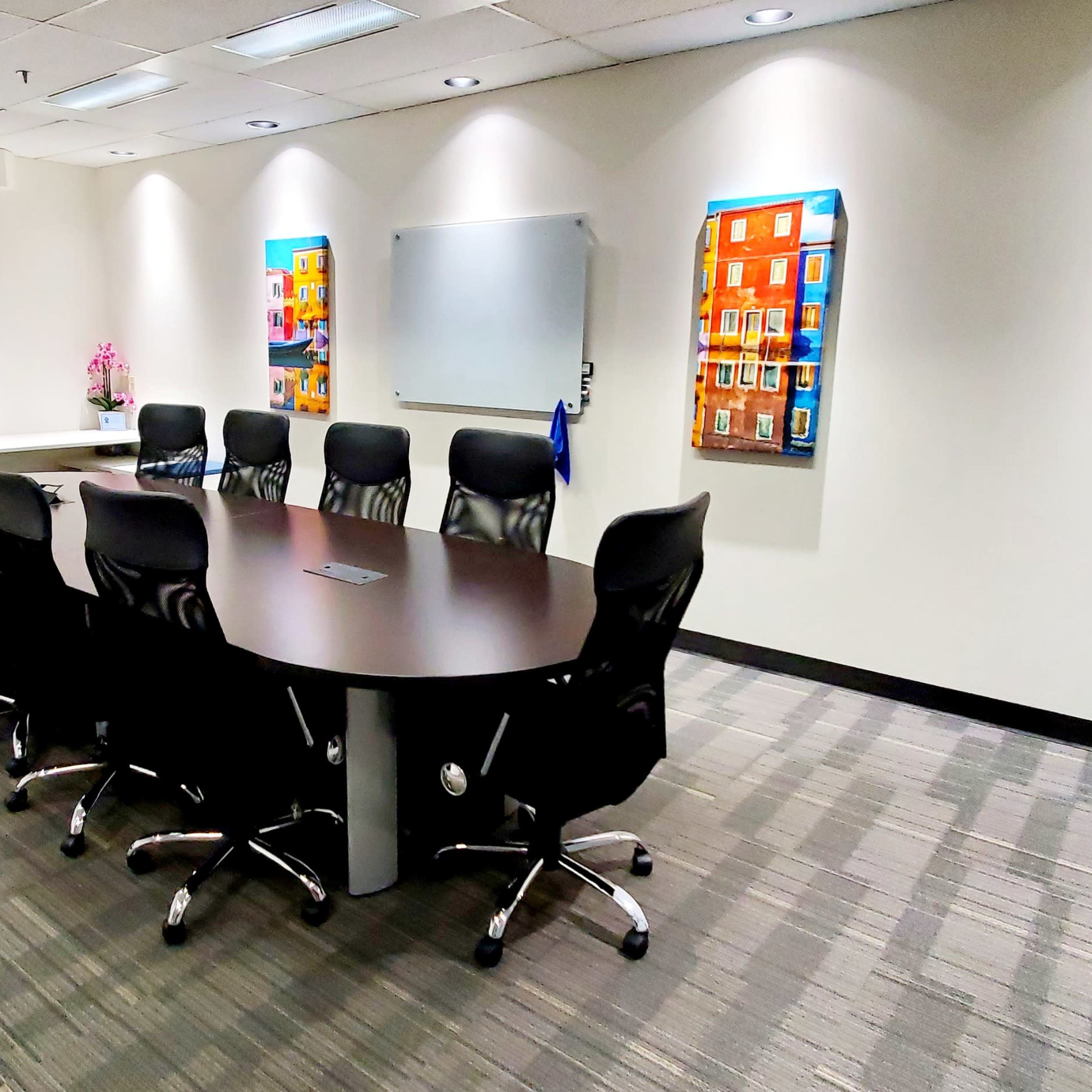 A conference room features a large oval table surrounded by nine black chairs, with a television screen and whiteboard on one wall and colorful artwork on another.