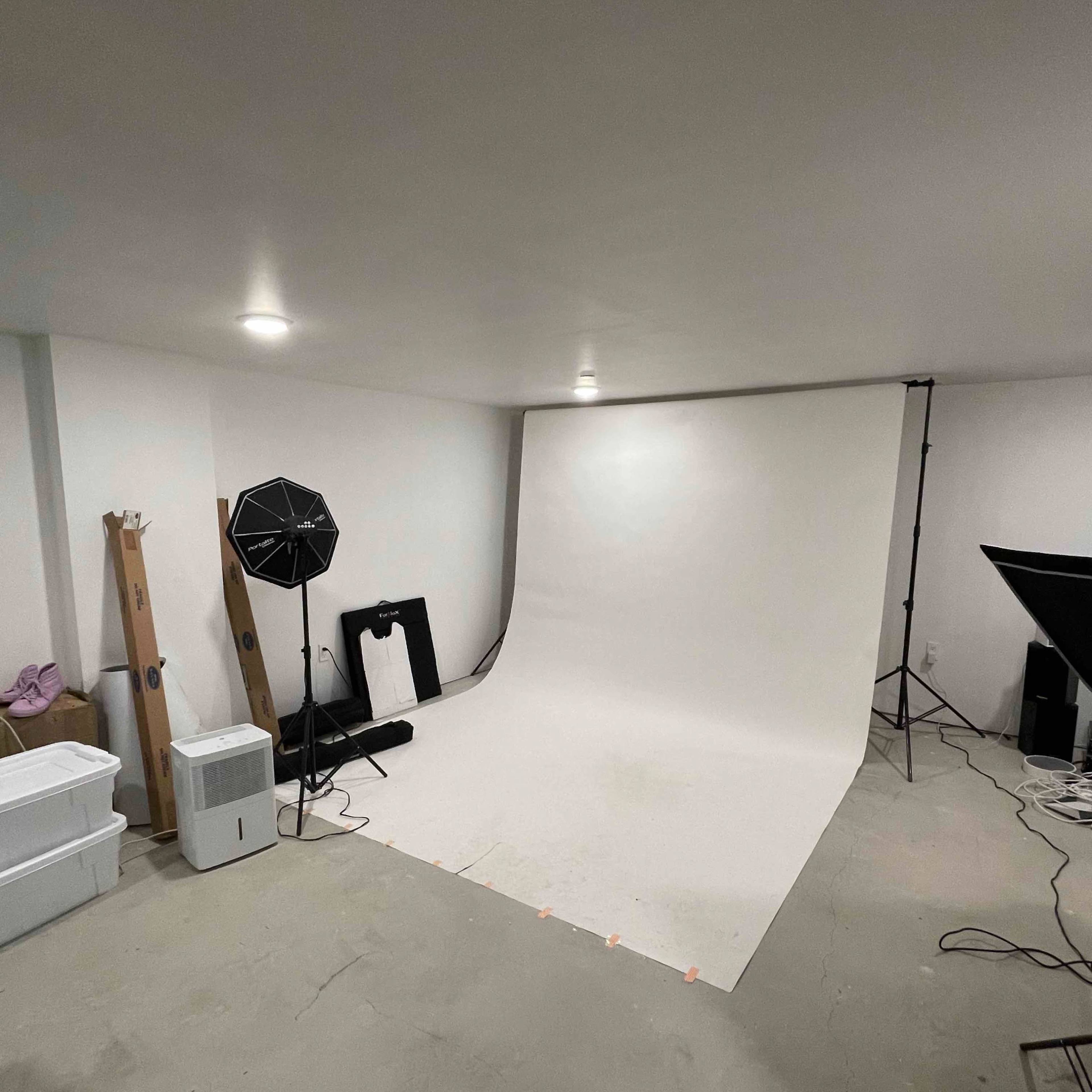 A photography studio setup with a seamless white backdrop, studio lights, and various props arranged against plain walls.