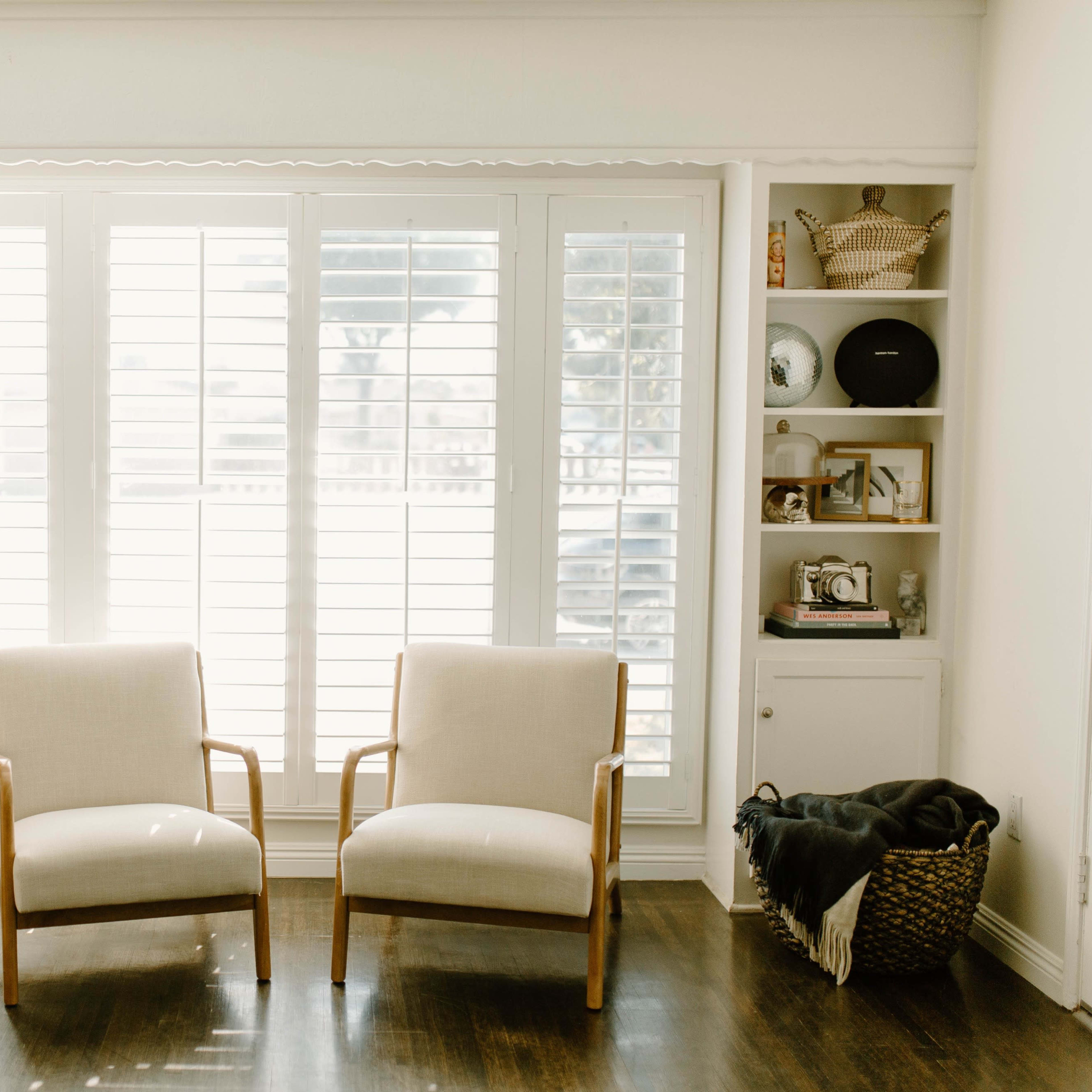 A living room with two cream-colored chairs facing a window covered by white shutters, flanked by built-in shelves displaying various decorative items.