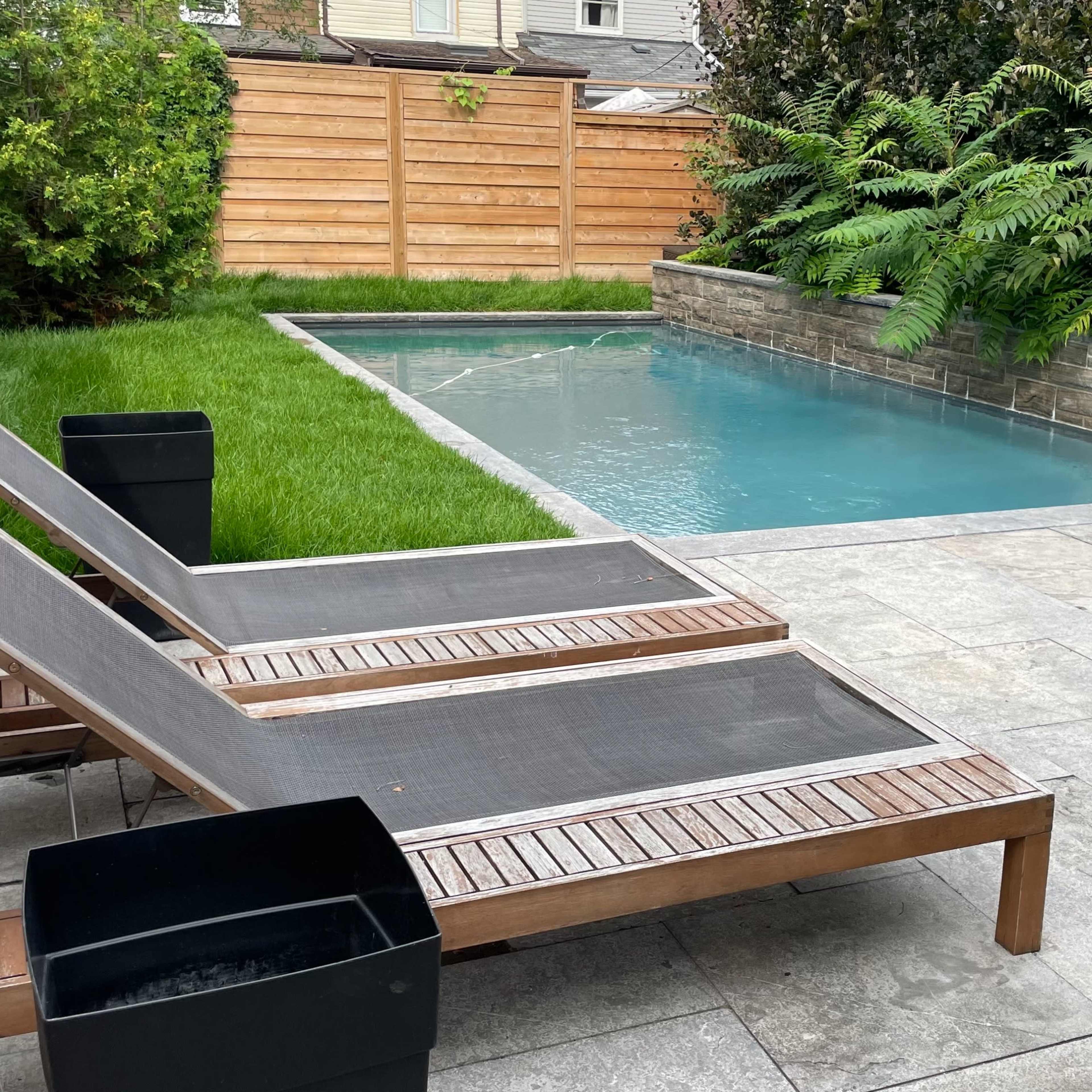 A small backyard features a swimming pool surrounded by grass and stone decking, with lounge chairs and a dining area set nearby.