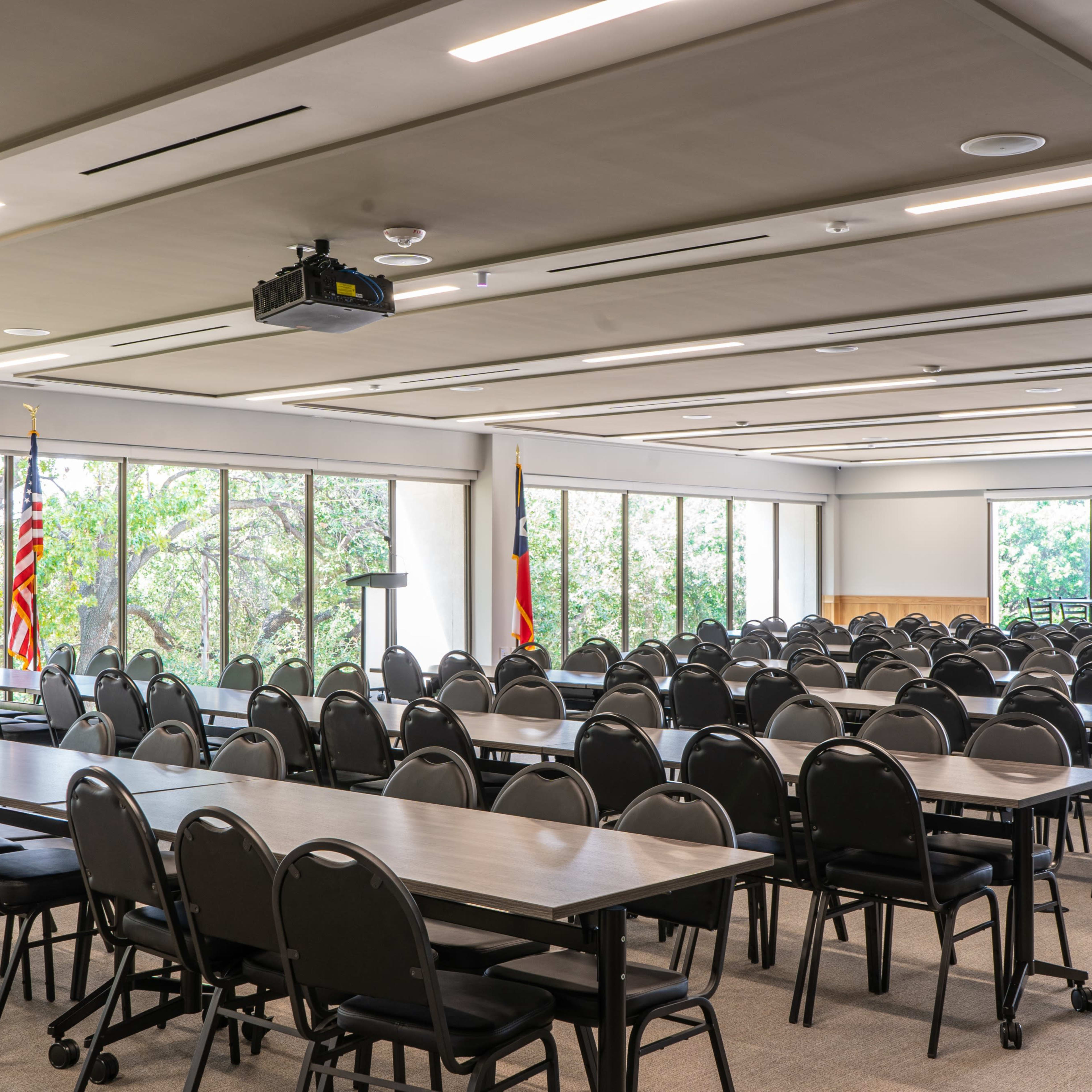 A large, empty conference room features multiple rows of tables and chairs, with American and state flags displayed near the windows.
