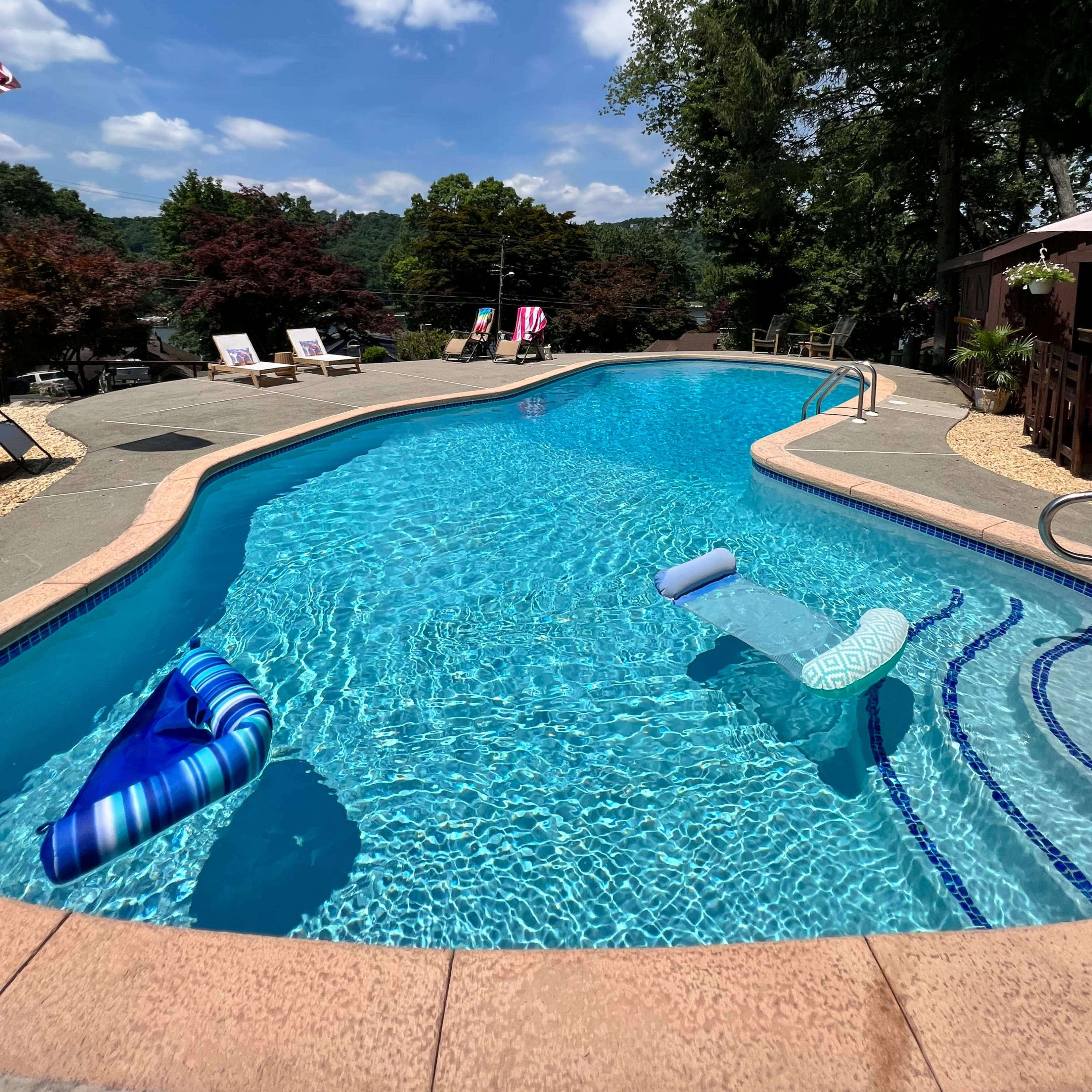 A clear swimming pool surrounded by trees and lounge chairs on a sunny day.