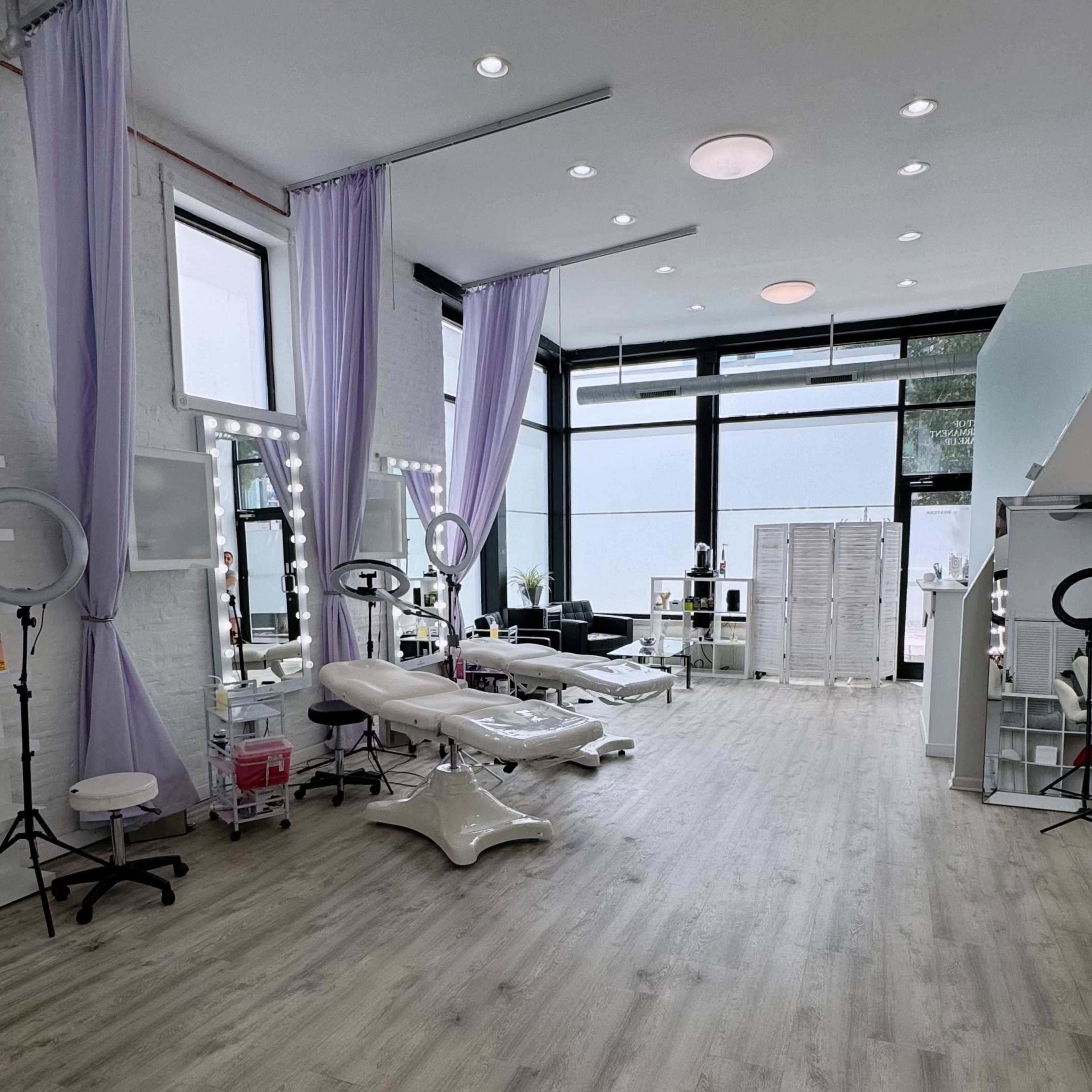 A spacious beauty studio with light-colored wooden flooring, large windows, and elegantly draped purple curtains.
