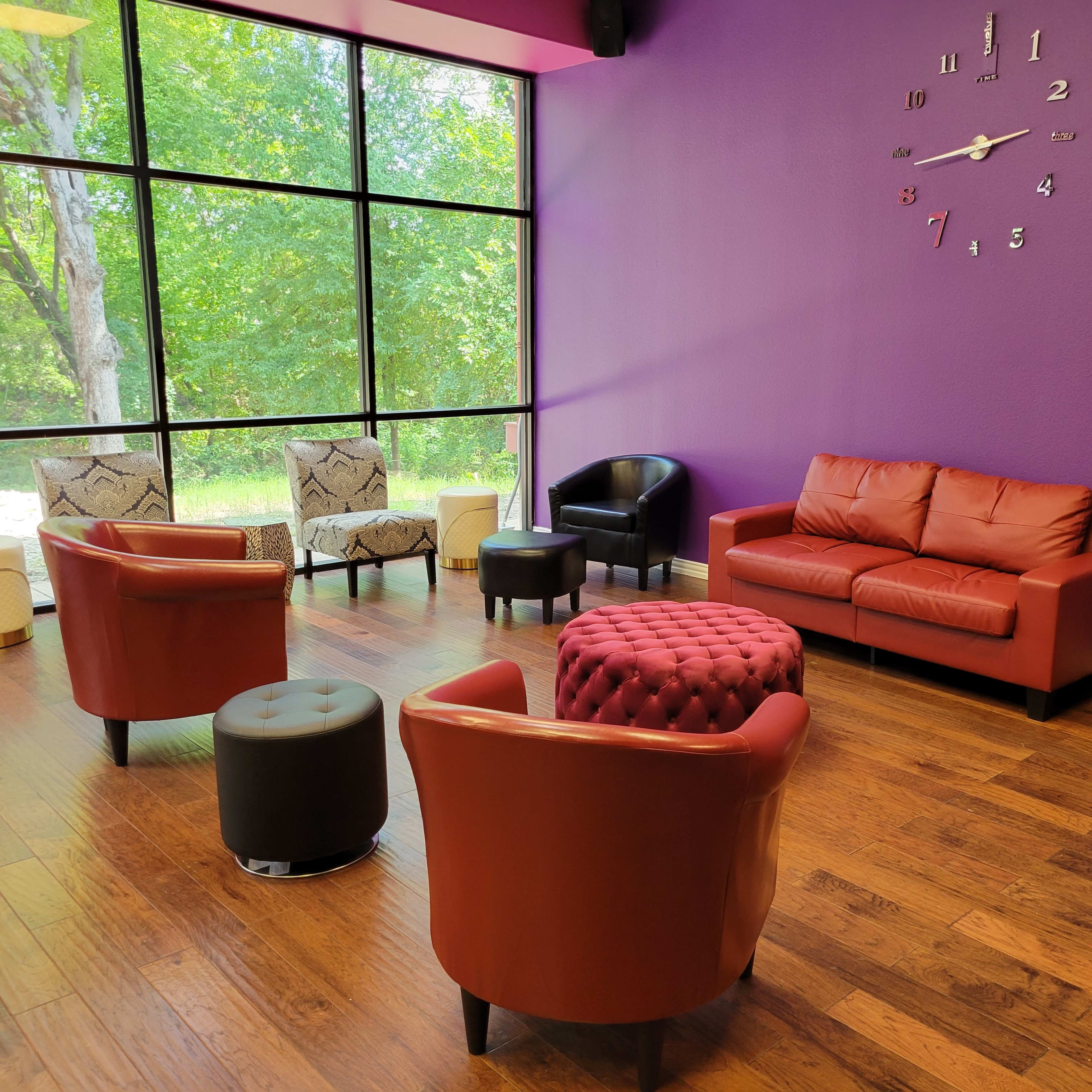 A brightly lit lounge area features a purple wall, a brown leather couch, and several seating options arranged around a central ottoman.
