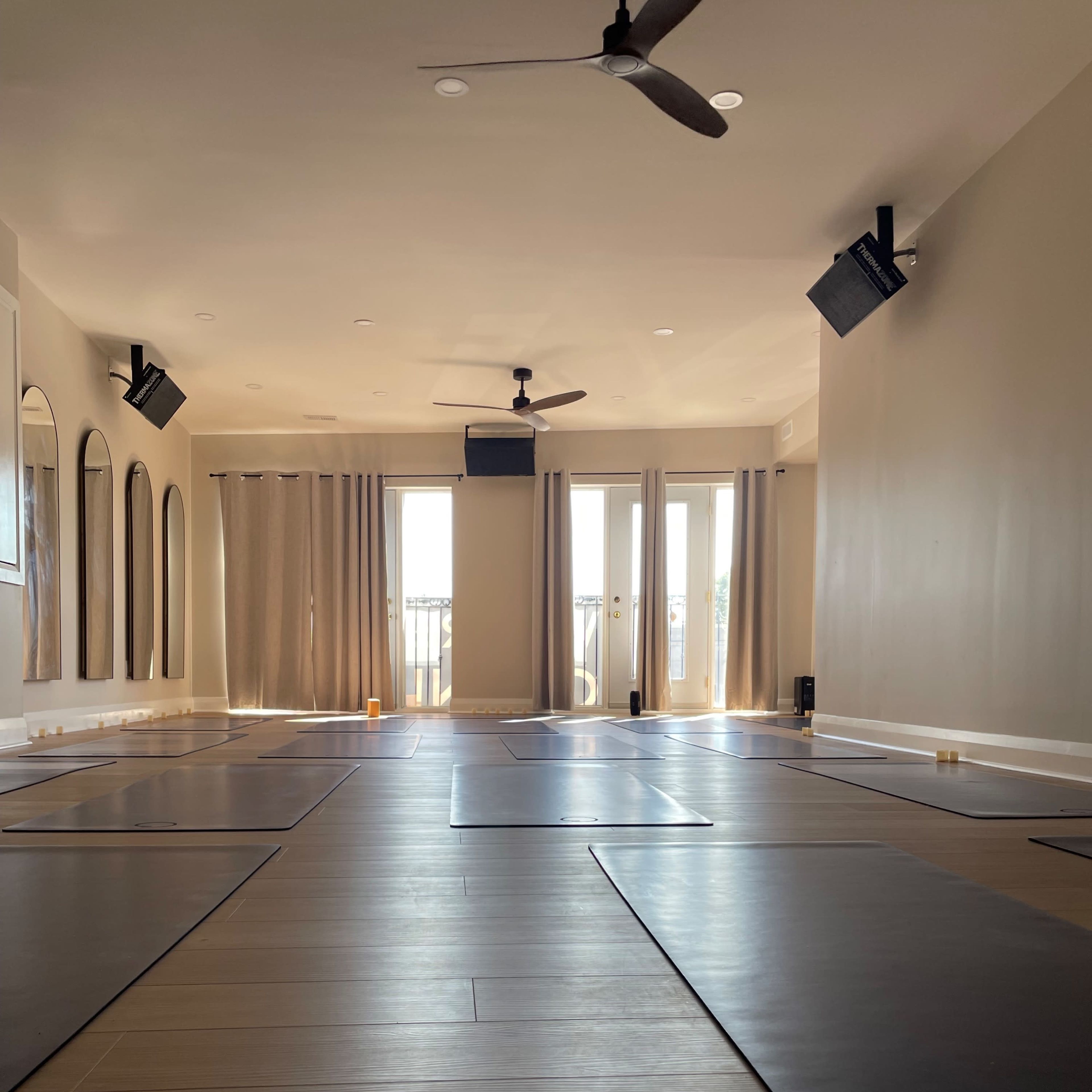 A spacious yoga studio with polished wooden floors, several yoga mats arranged on the ground, and large windows covered by light curtains.
