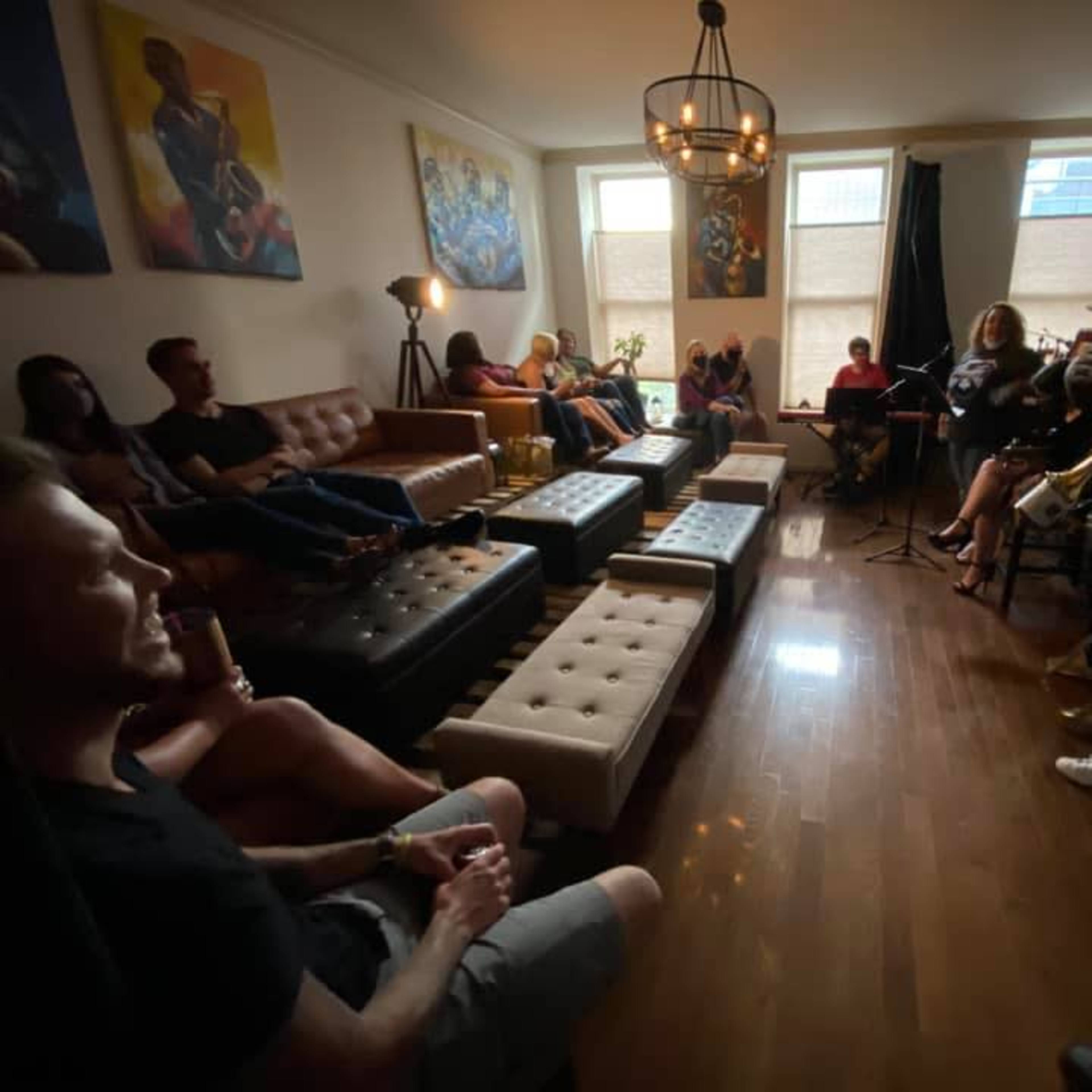 A group of people are seated in a cozy living room while a small band performs music in the corner.