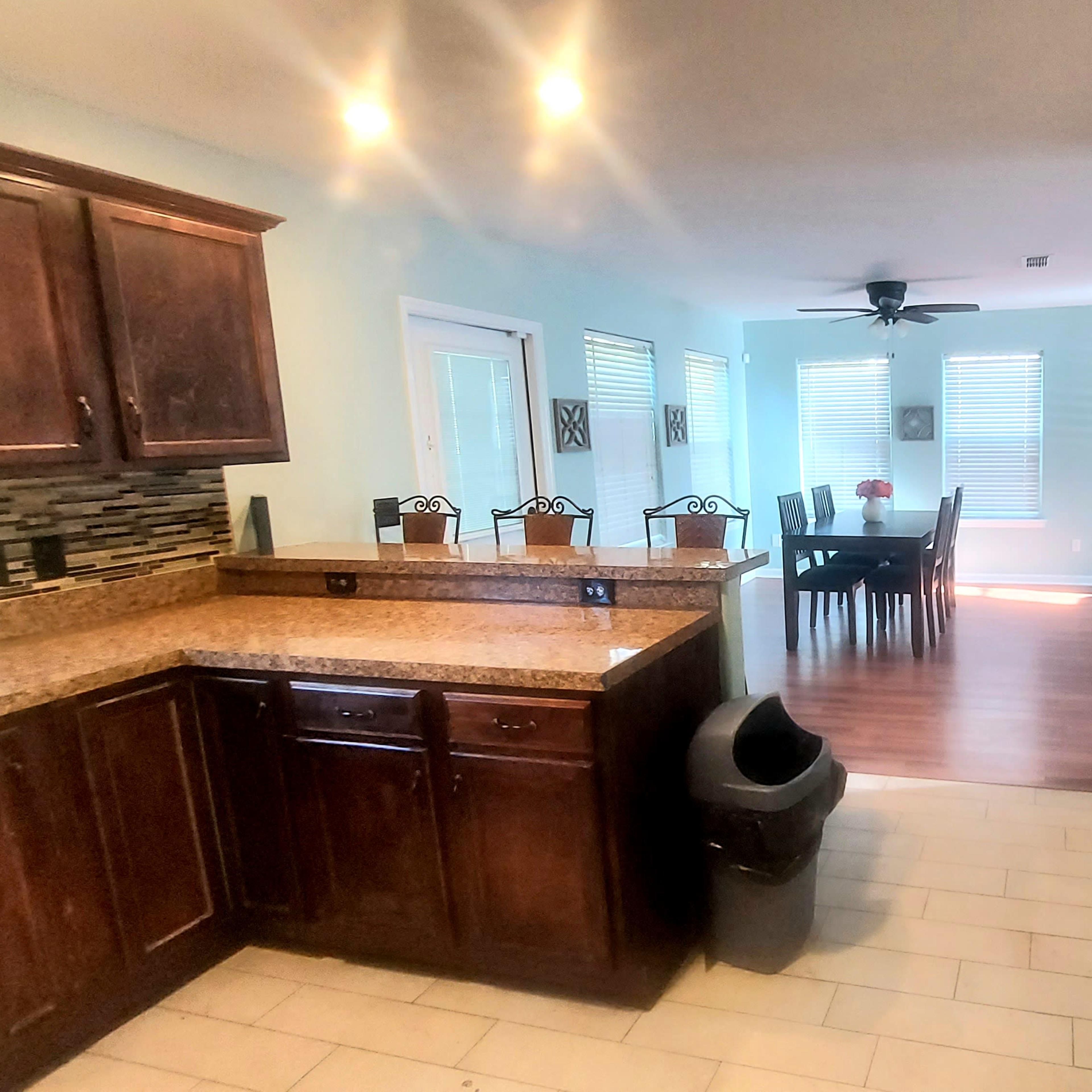 A kitchen area with brown cabinets and a granite countertop, leading into a dining space with a table and chairs under a ceiling fan.