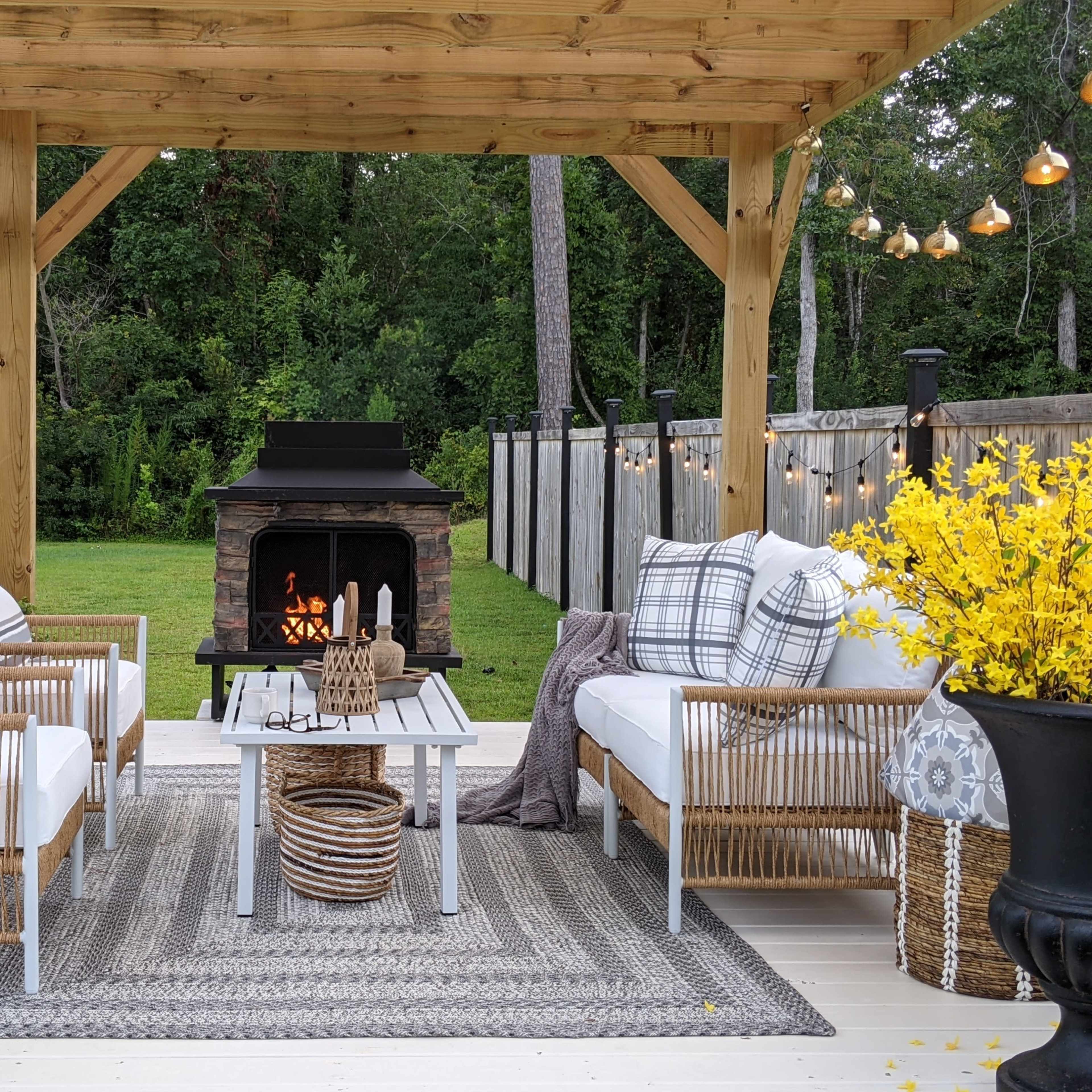 The scene features a cozy outdoor seating area under a wooden pergola, furnished with white armchairs and a table, with a stone fire pit in the background surrounded by greenery.